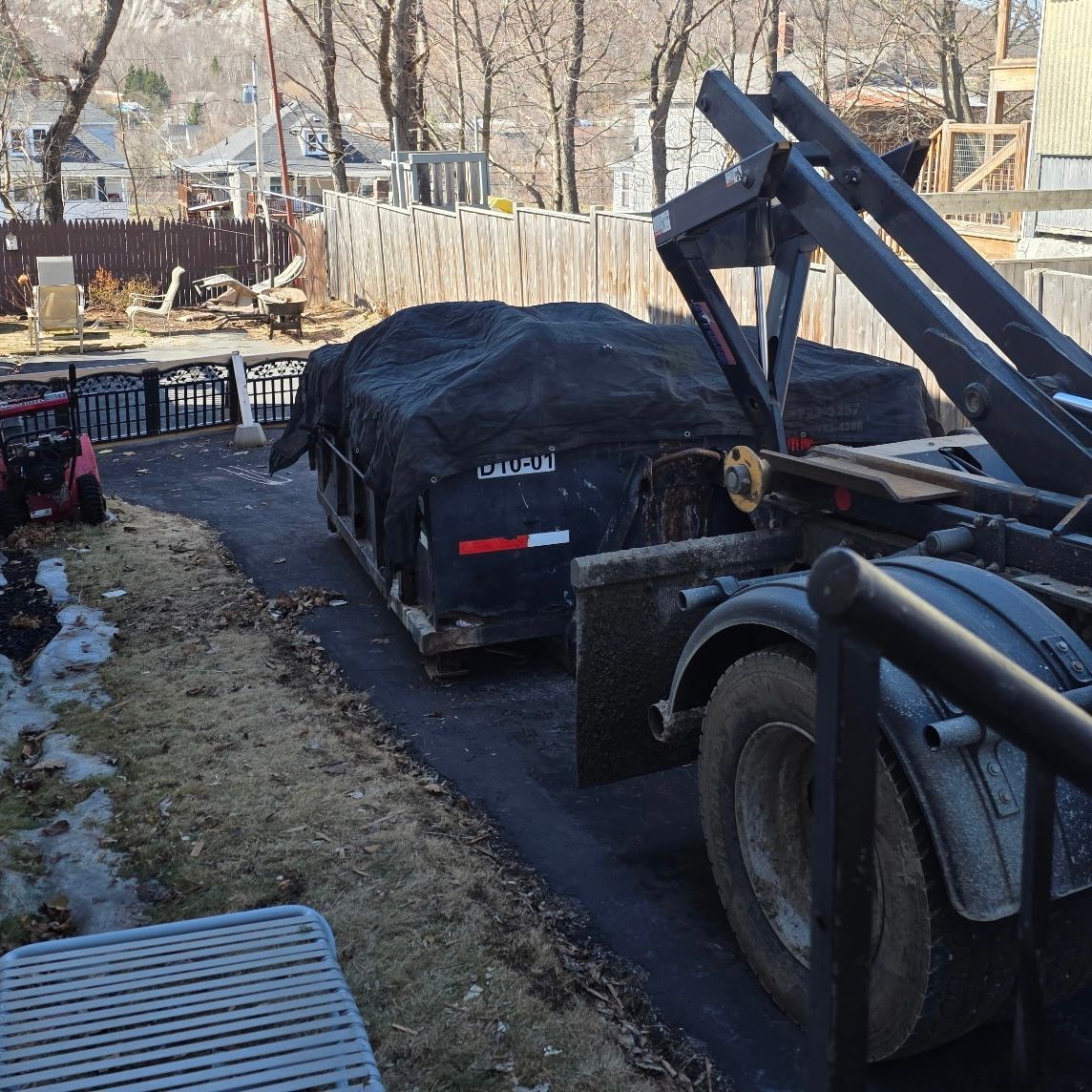 A black dumpster covered with a tarp, being loaded by a truck in a residential driveway.