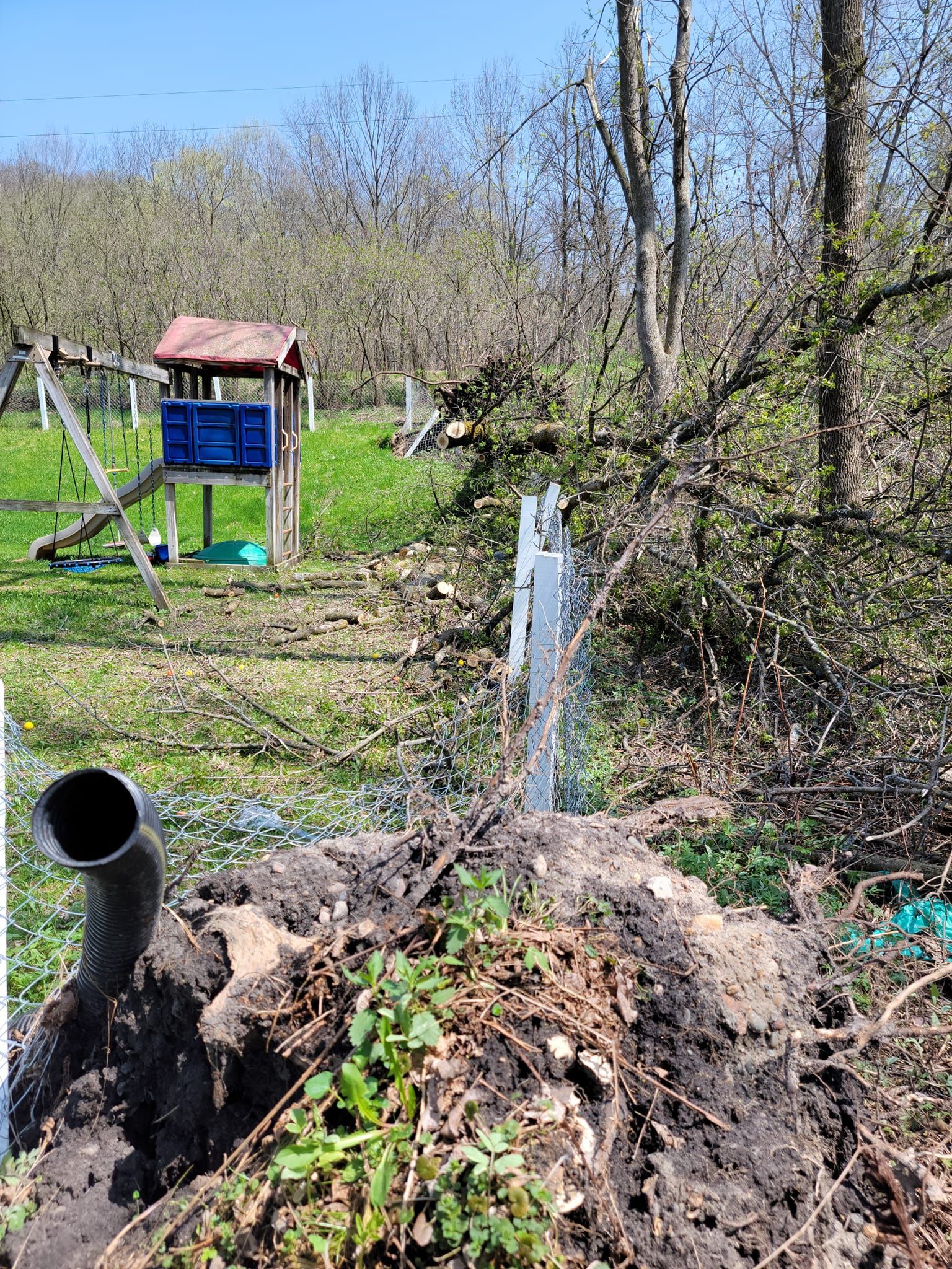 A water pipe gushing water near dirt and foliage, with a playhouse and trees in the background.