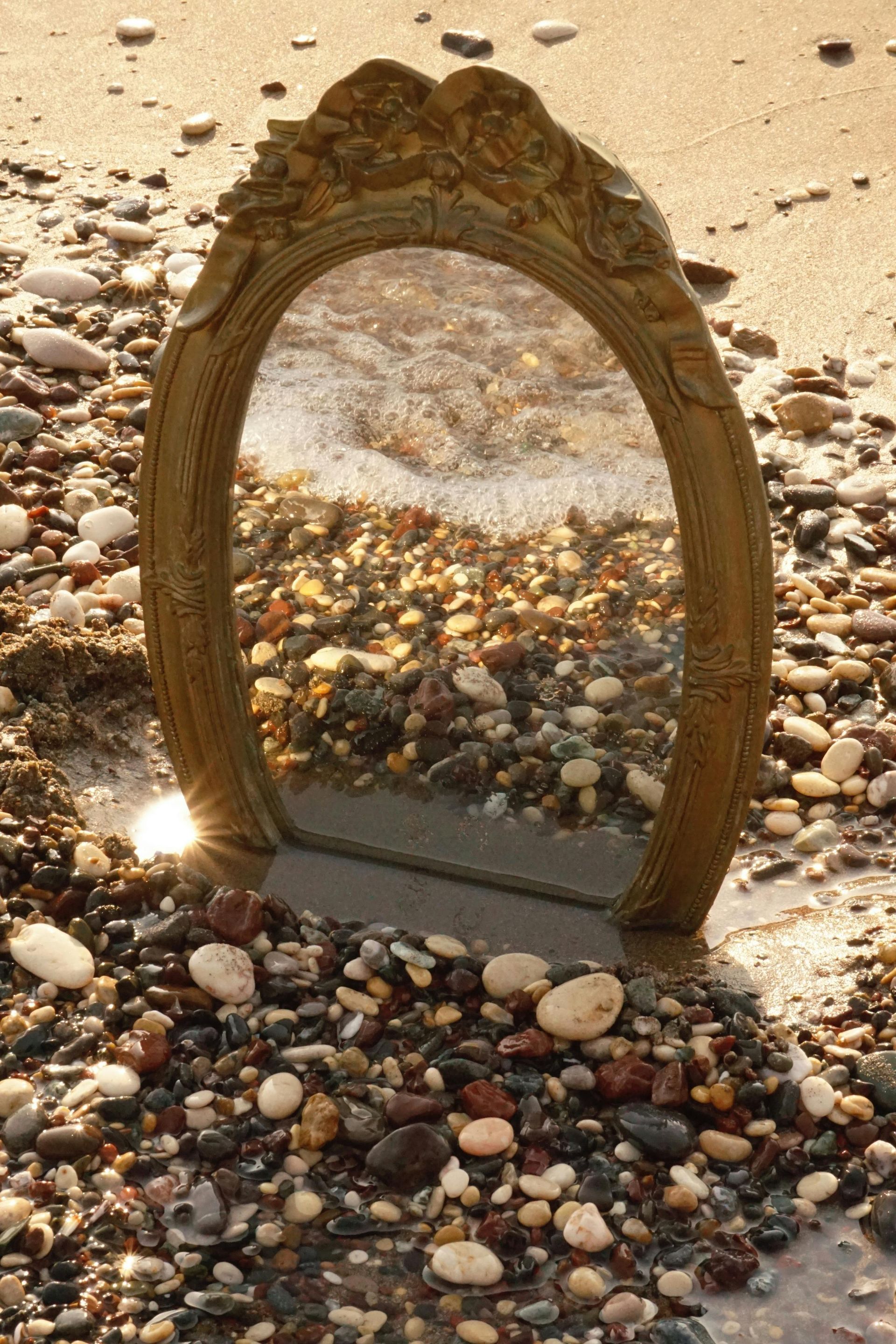 An ornate, oval-framed mirror stands upright on a pebble-covered beach, reflecting the surrounding stones and sea foam.