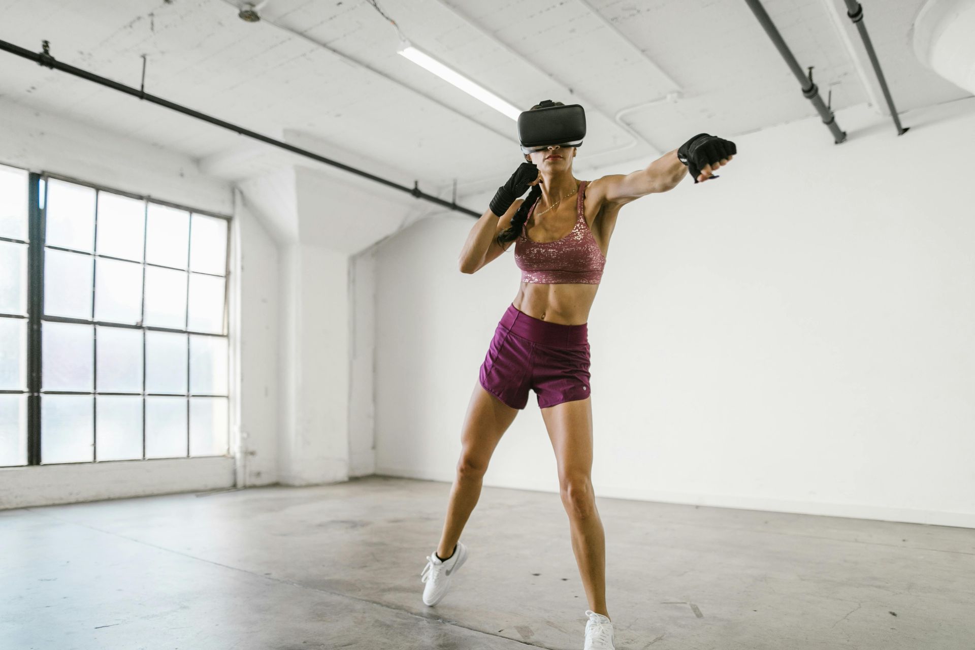 An athlete in a workout top and shorts wears a VR headset while practicing boxing in a bright, industrial studio space.
