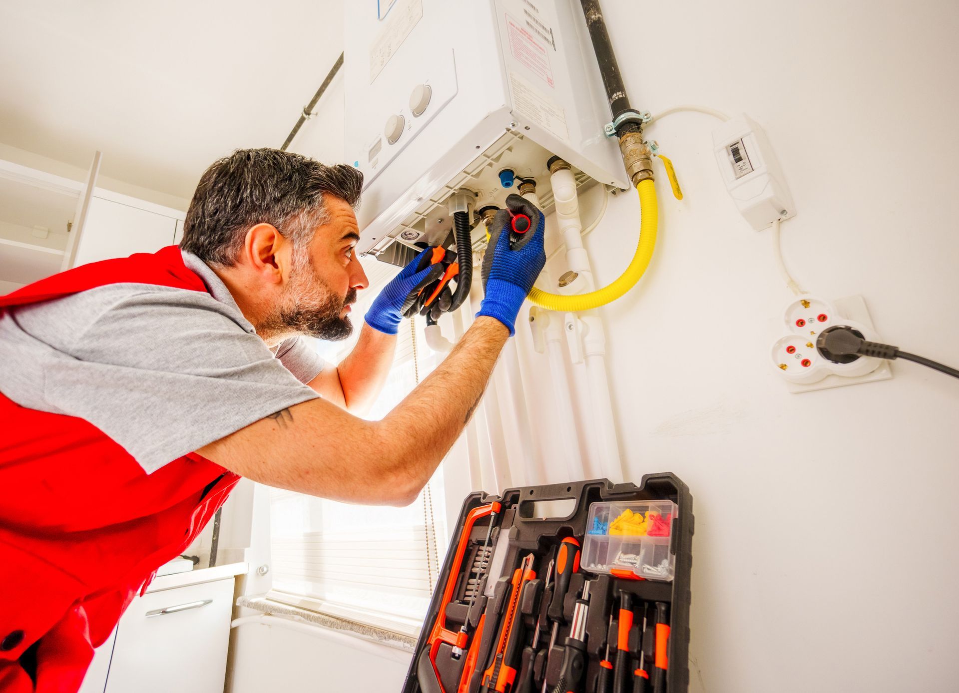 Plumber in red vest fixing a gas boiler with tools, blue gloves on, in a white utility room.