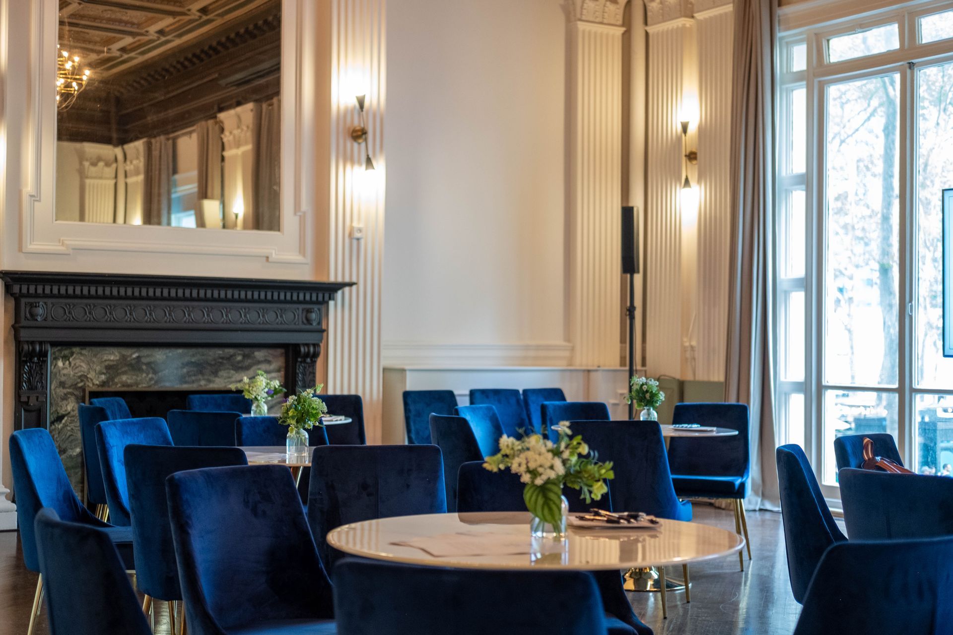 Elegant dining room with blue velvet chairs, round tables, a black fireplace, and tall windows
