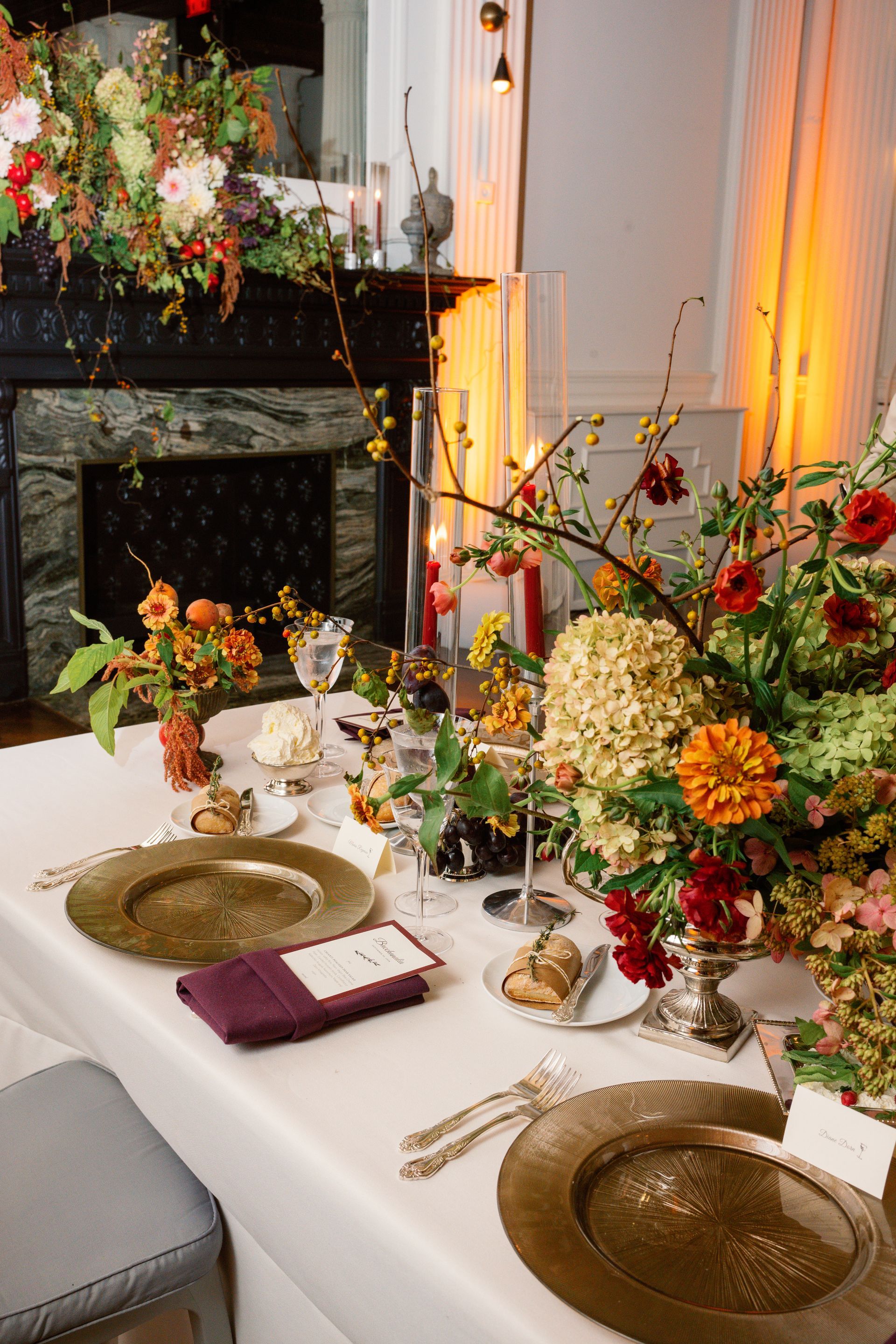 Elegant dining table with white tablecloth, gold plates, and colorful floral centerpieces by a fireplace