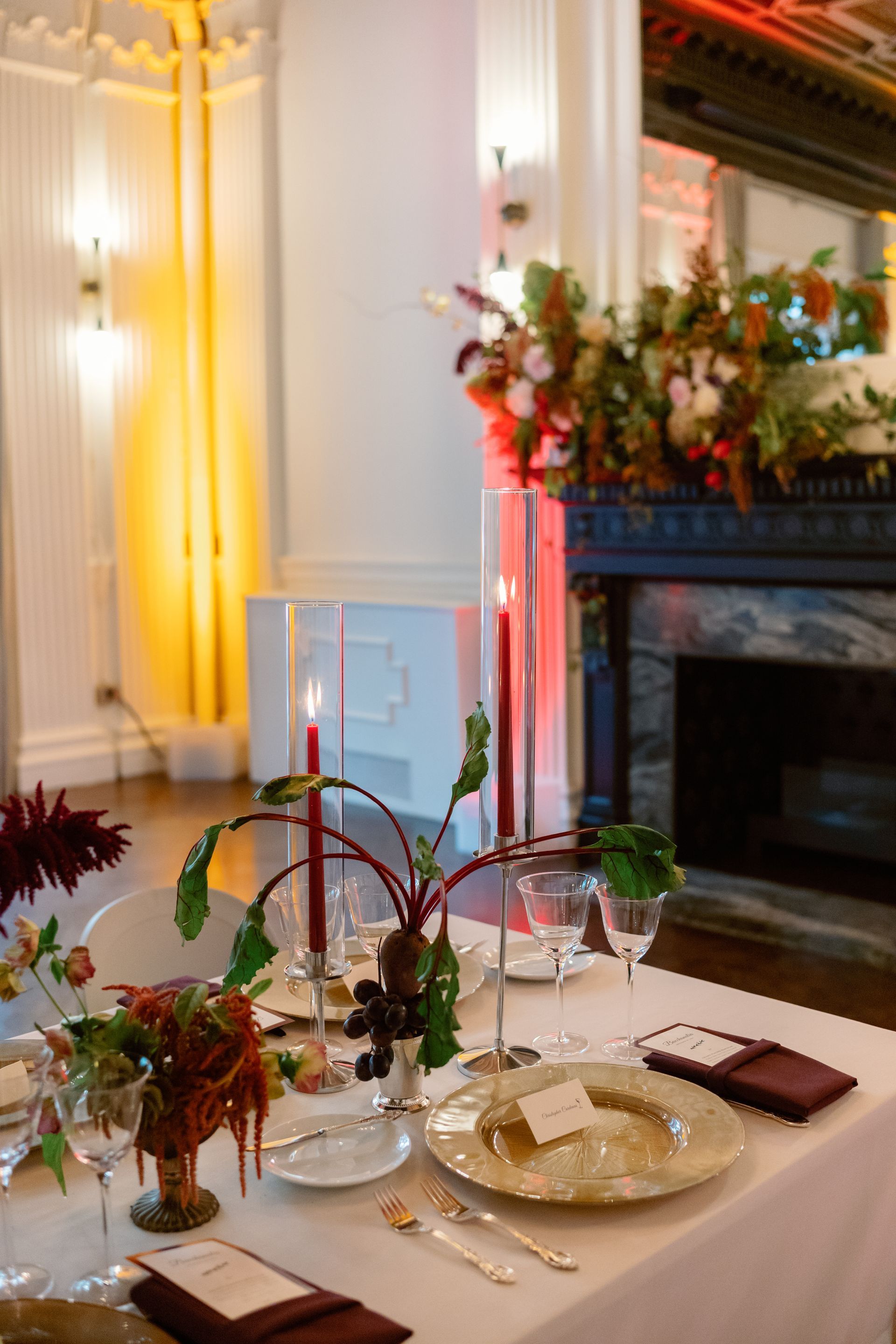 Elegant dining table set with candles, flowers, and glassware beside a fireplace in a warmly lit room