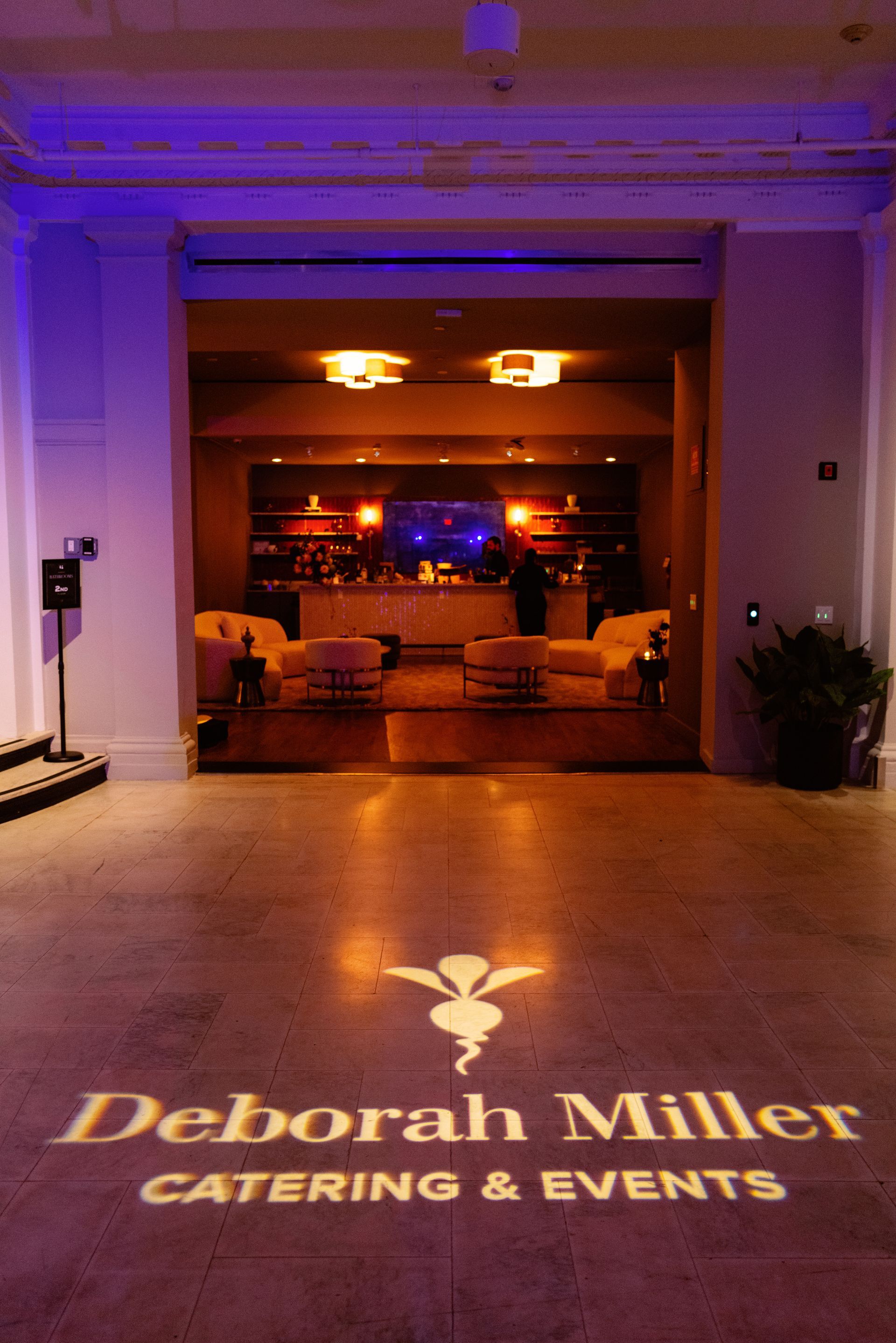 Deborah Miller Catering & Events logo projected on a hotel lobby floor with a warmly lit event space beyond