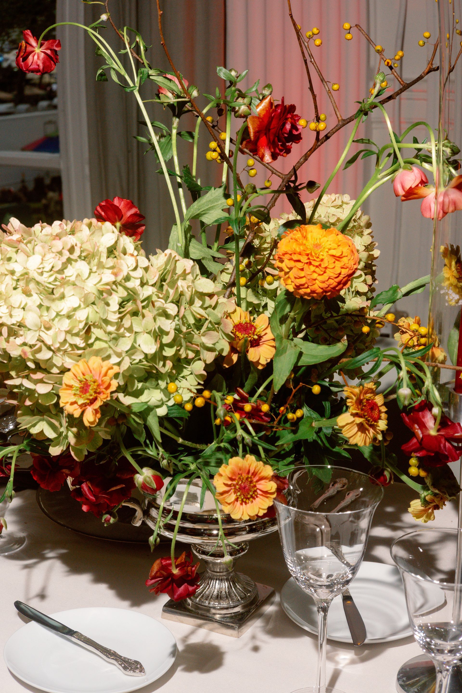 Floral centerpiece with white, orange, and red flowers arranged on a dining table with glassware and plates