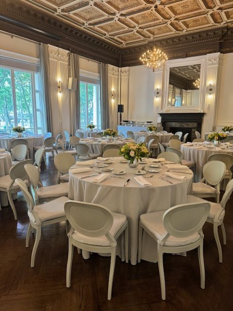 Elegant dining room with round tables, white chairs, chandeliers, and a fireplace beneath ornate ceiling moldings