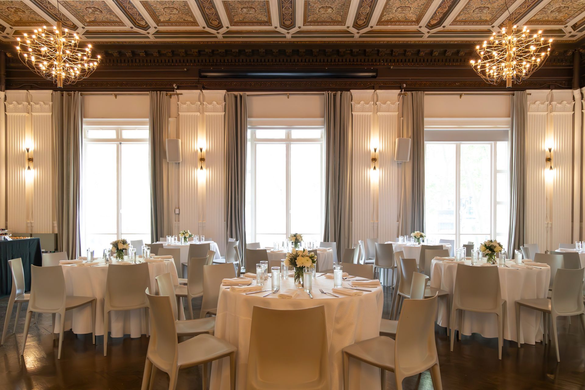 Elegant banquet hall with round tables, white chairs, chandeliers, and tall windows in warm light