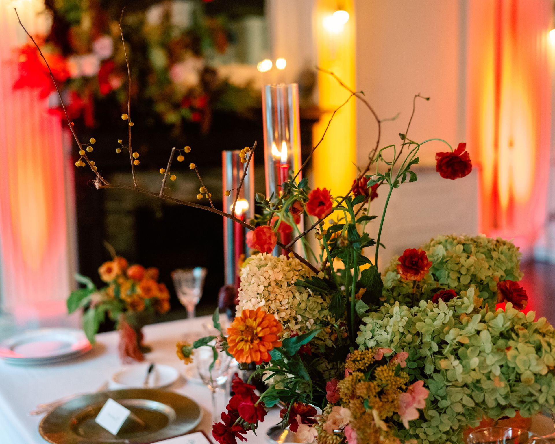 Elegant table centerpiece with red, orange, and cream flowers beside lit candles on a formal dining table