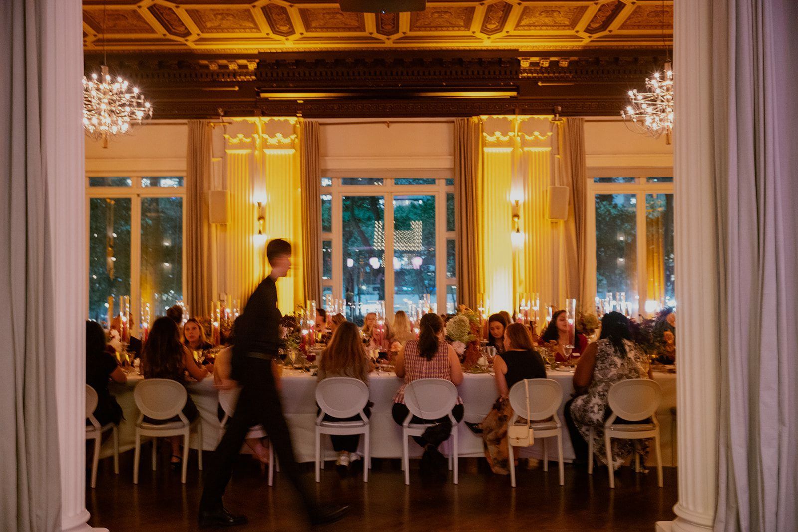 A waiter walks past a long table set for a formal dinner, with guests seated in a warmly lit elegant room.
