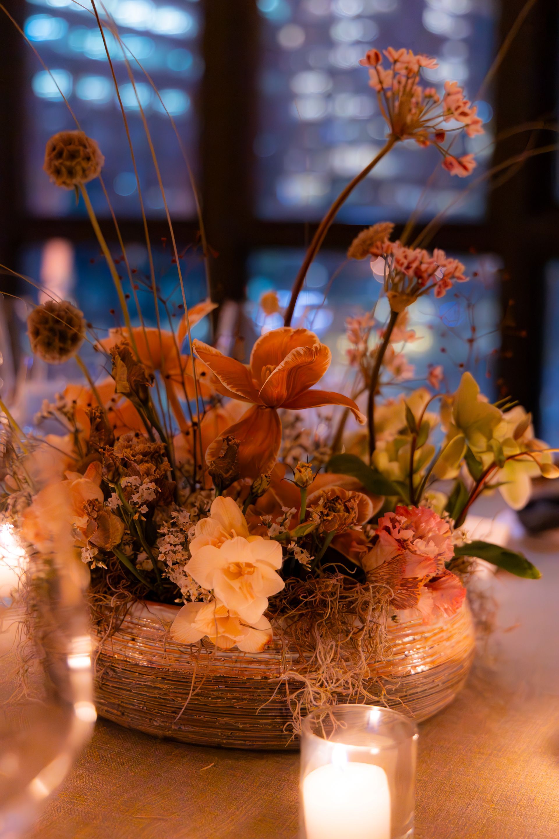 Orange and white floral arrangement in a woven bowl with candles glowing on a table at dusk