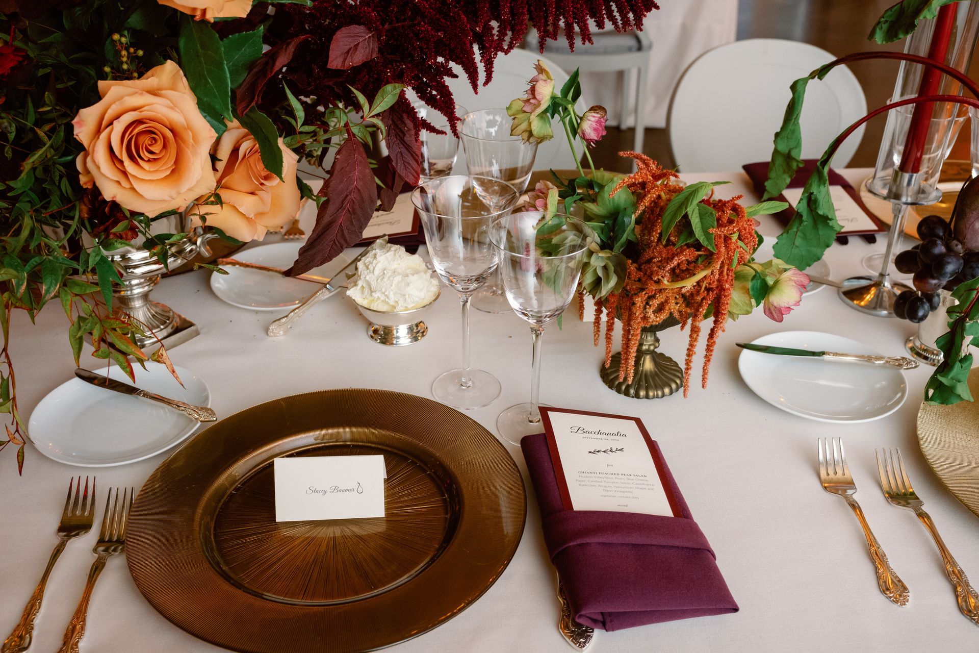 Elegant table setting with gold plates, burgundy napkins, roses, and glassware on a white tablecloth