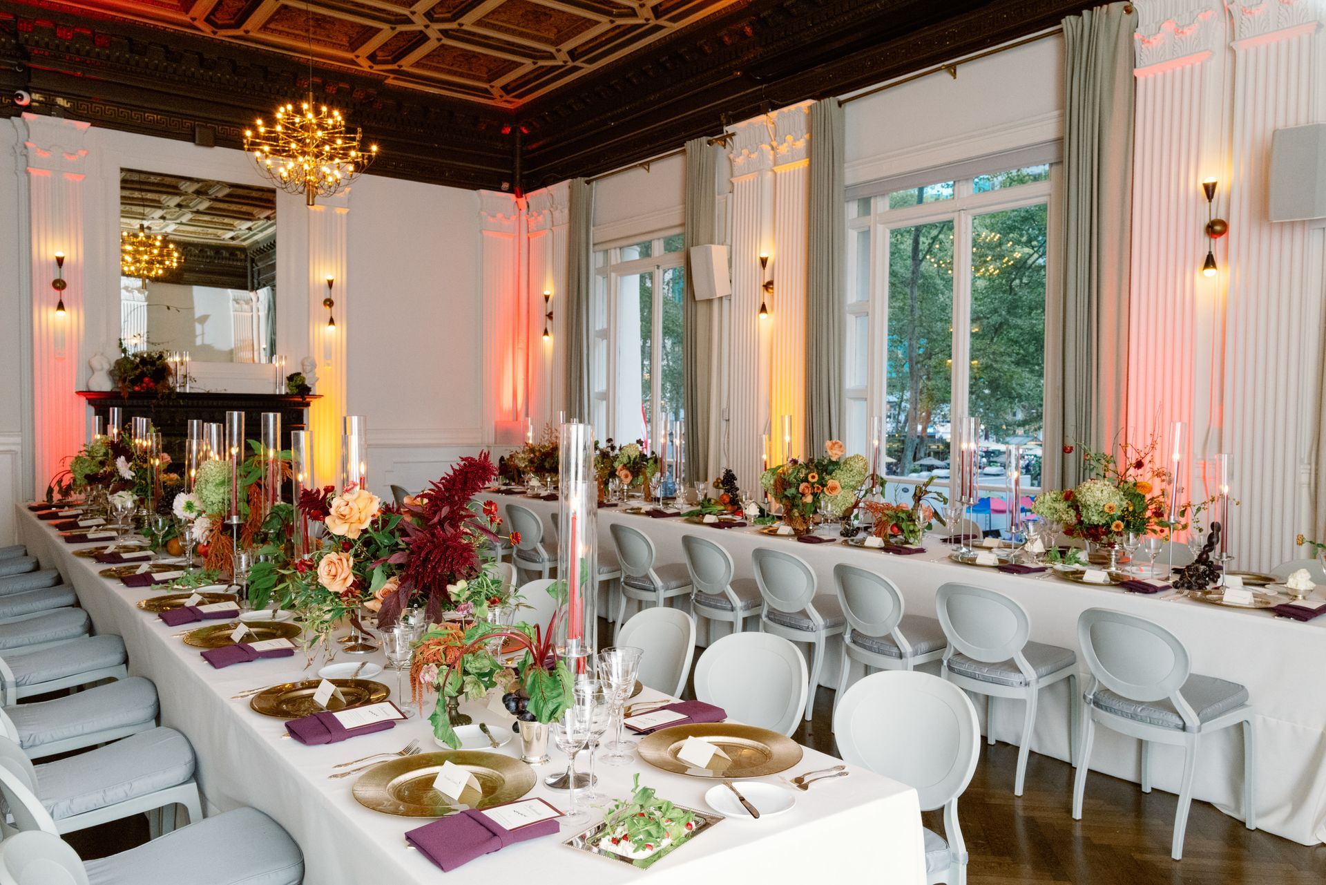 Elegant dining room with long white banquet tables, floral centerpieces, and tall windows with soft pink lighting