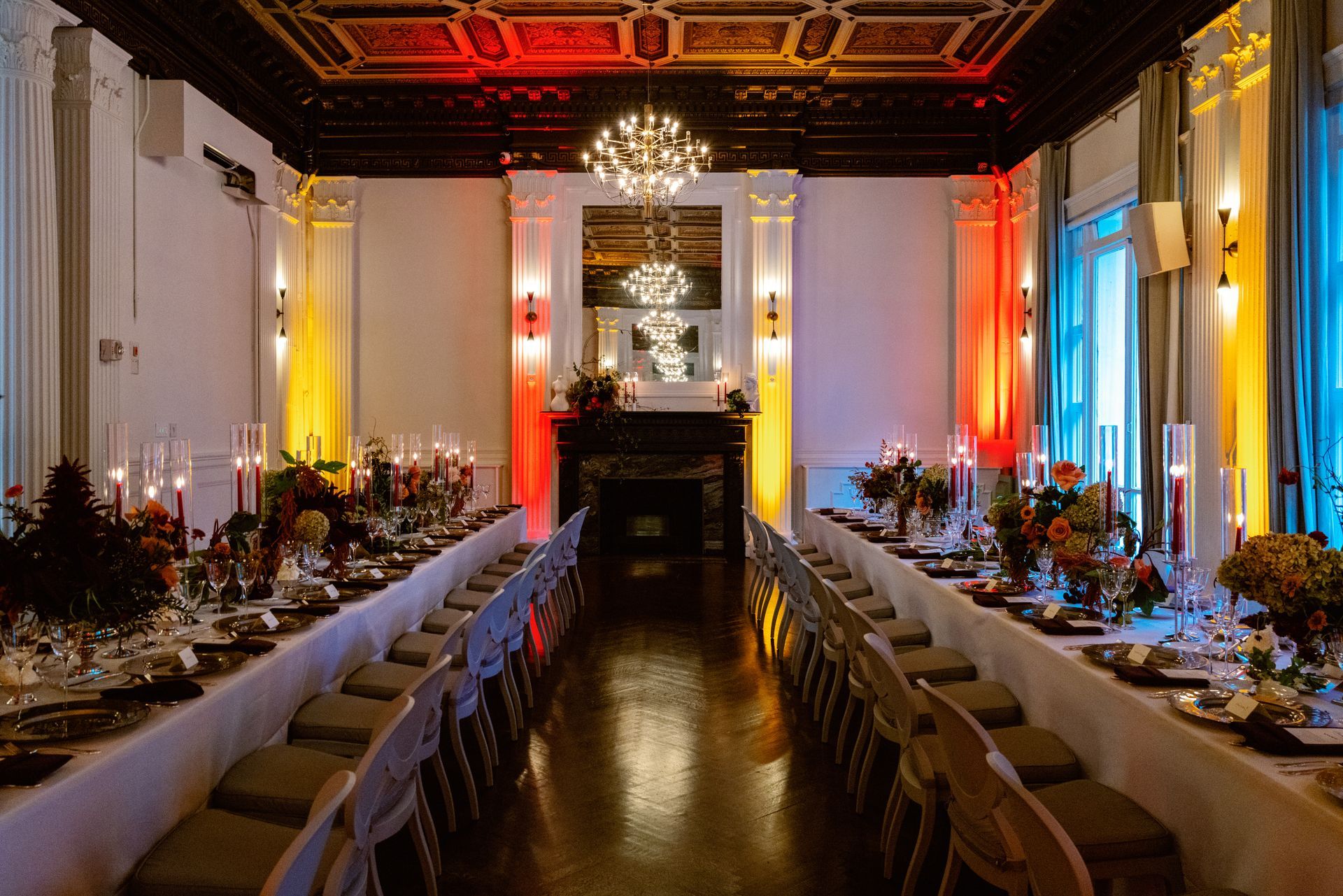 Elegant banquet hall with long decorated tables, chandeliers, and colorful uplighting