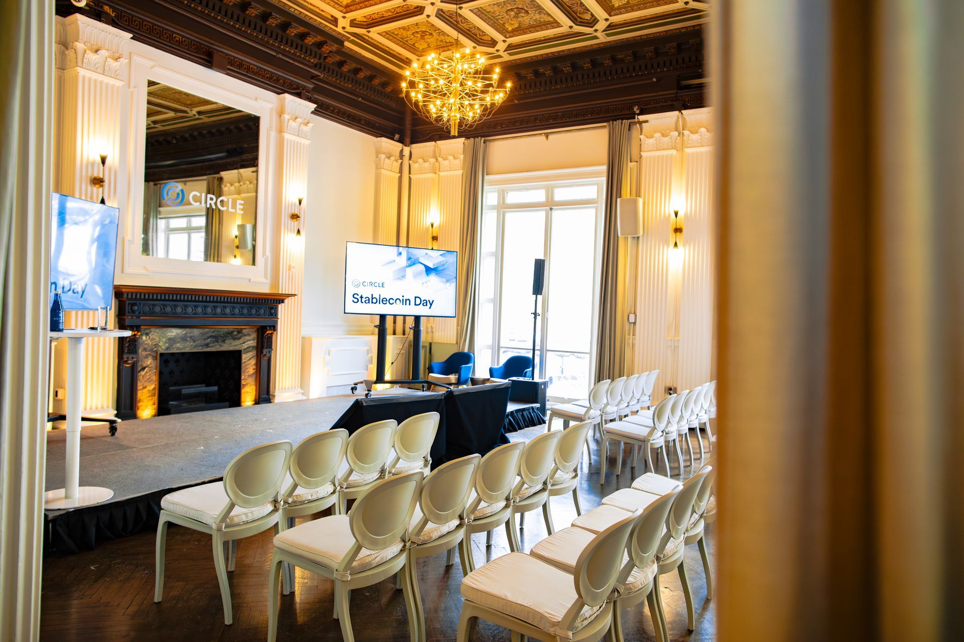 Elegant conference room with chandeliers, rows of white chairs, and screens beside a fireplace stage
