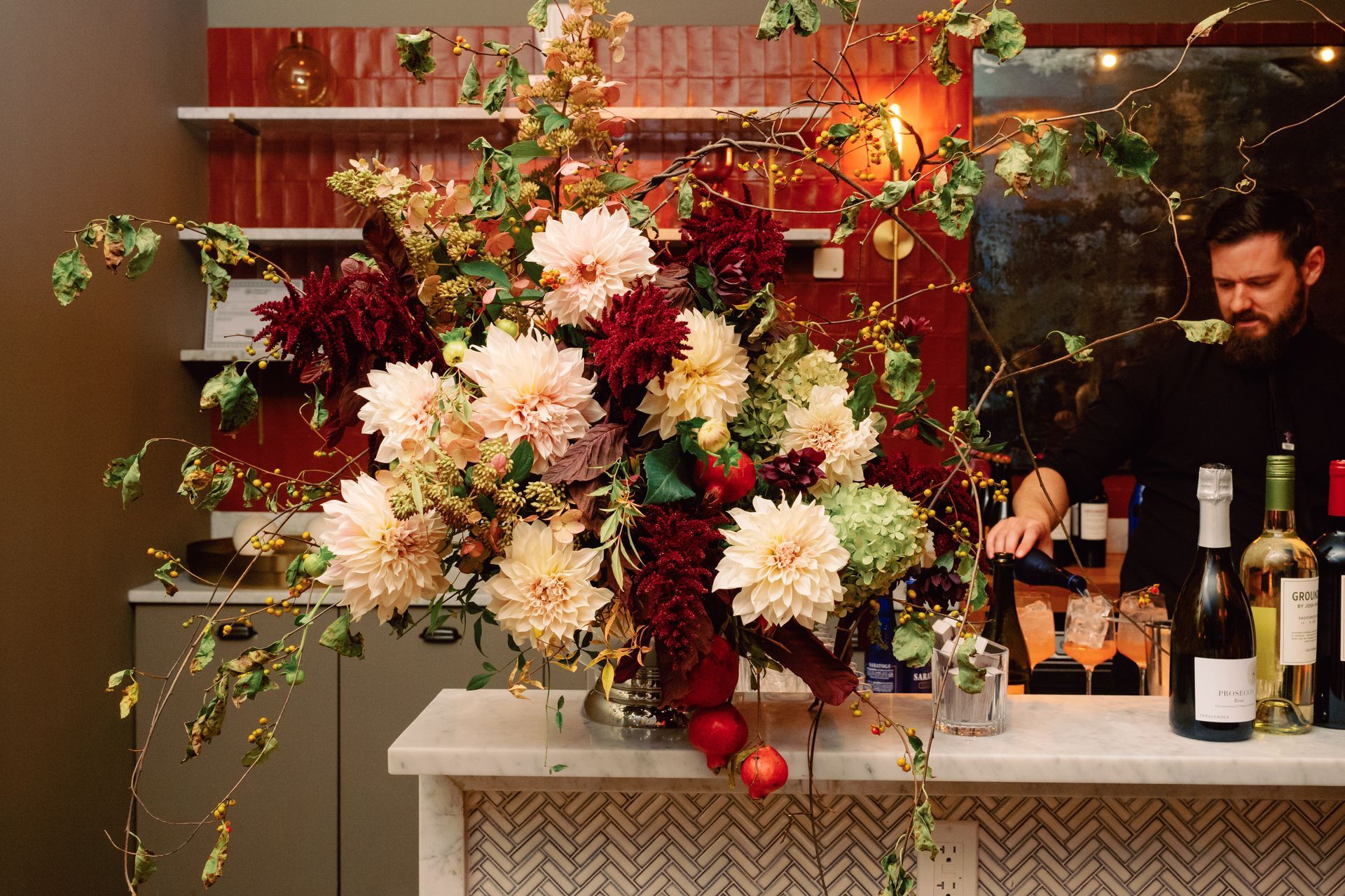 Large floral arrangement on a bar counter with bottles and a bartender in a warm, dimly lit setting