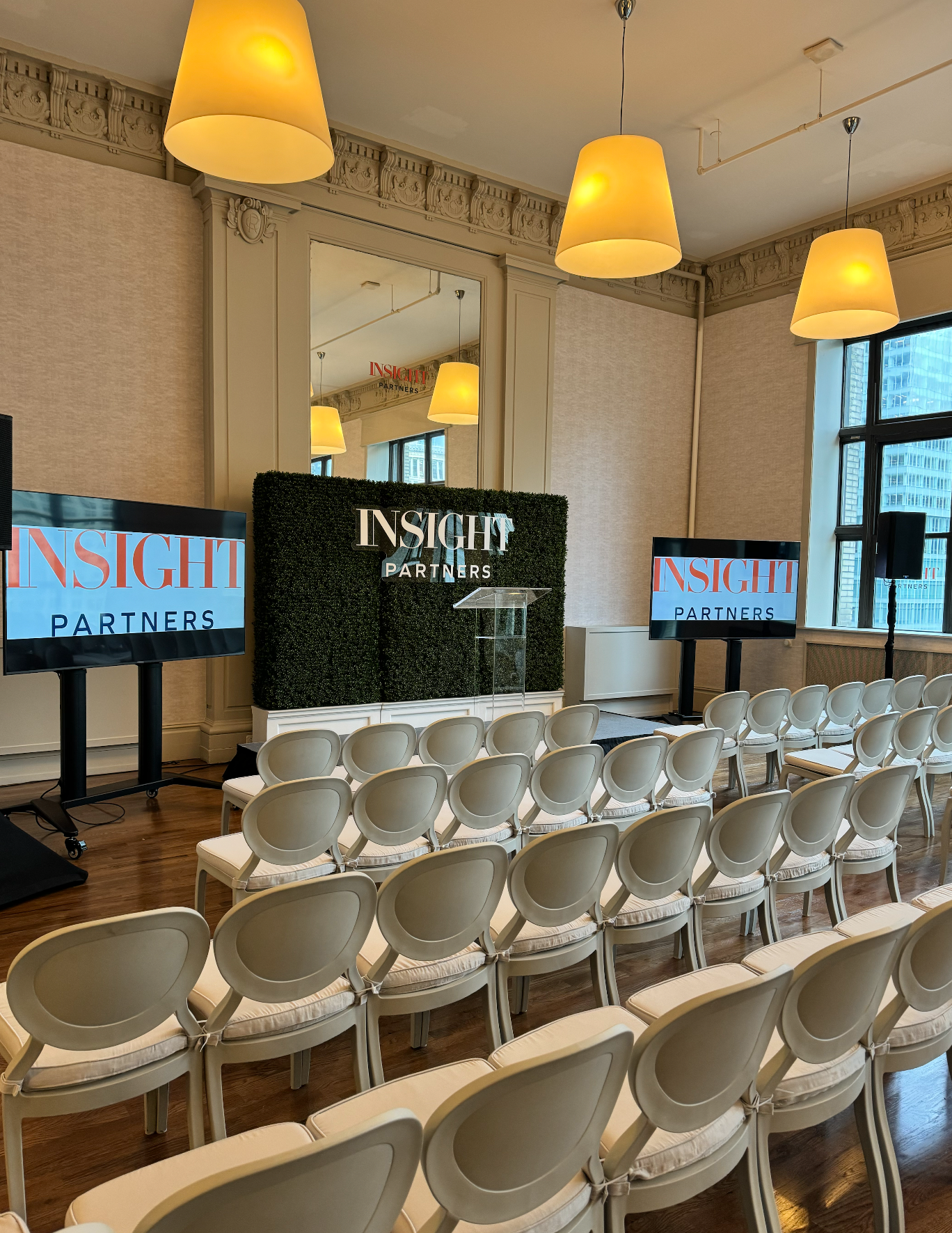 Conference room with rows of white chairs facing a stage and large screens under warm hanging lights