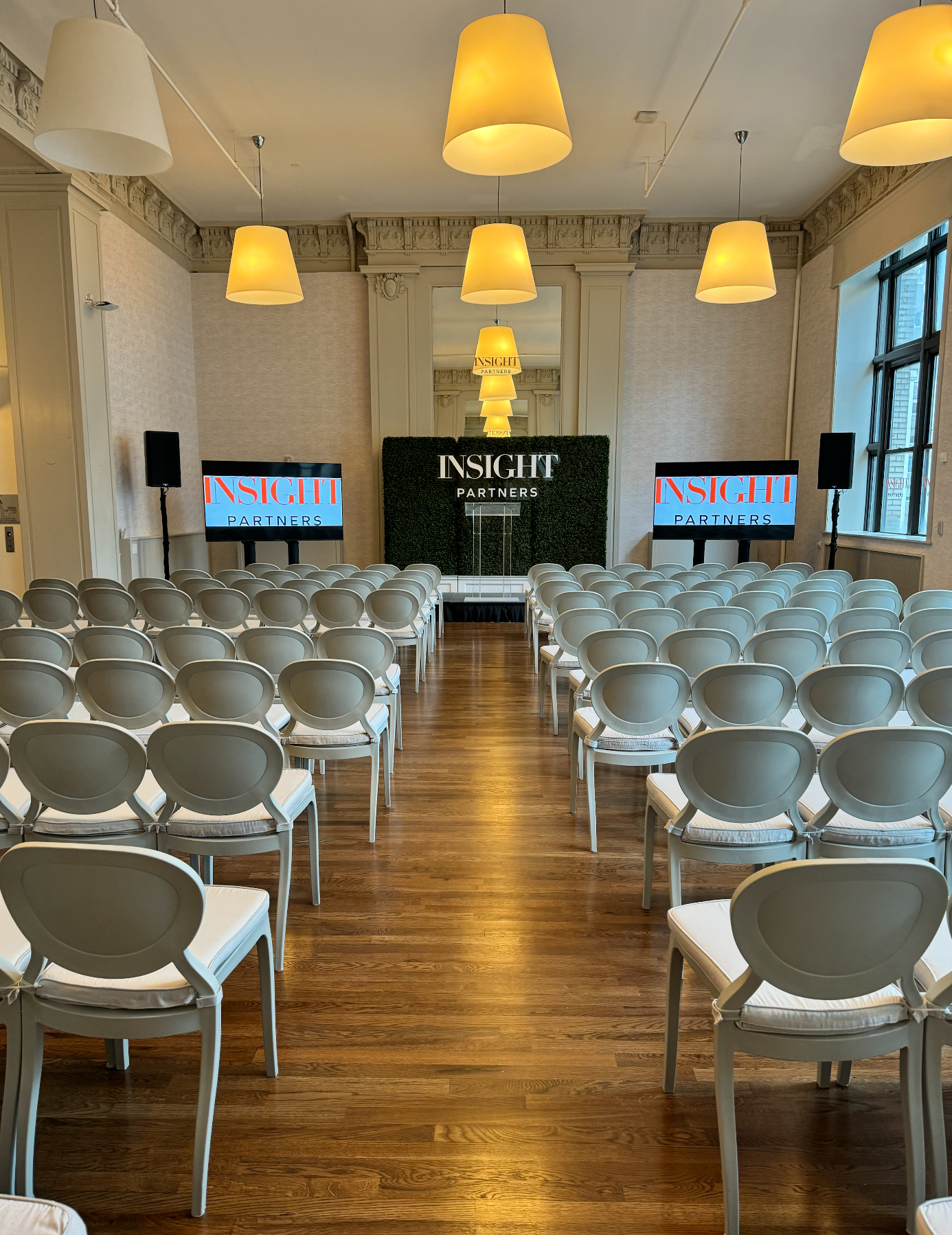 Conference room with rows of white chairs facing a podium and screen under warm hanging lights.