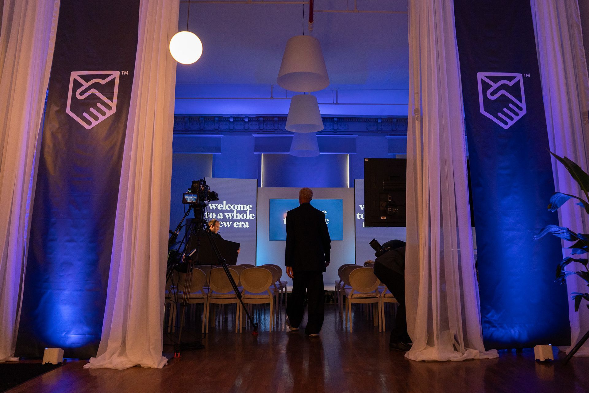 Blue-lit event hall with draped entrance, chairs, and a person walking toward a projection screen