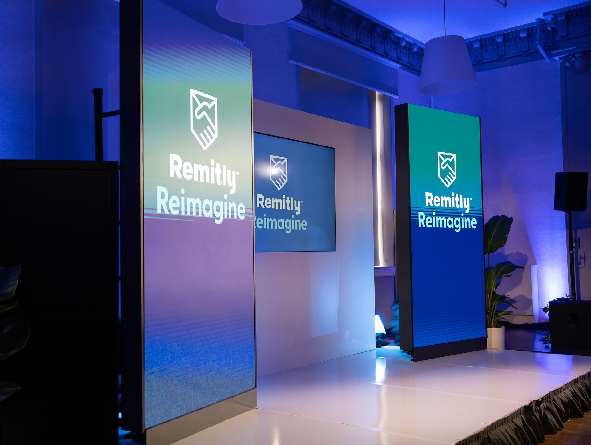 Blue-lit event stage with tall screens displaying the “Remitly Reimagine” logo and text.