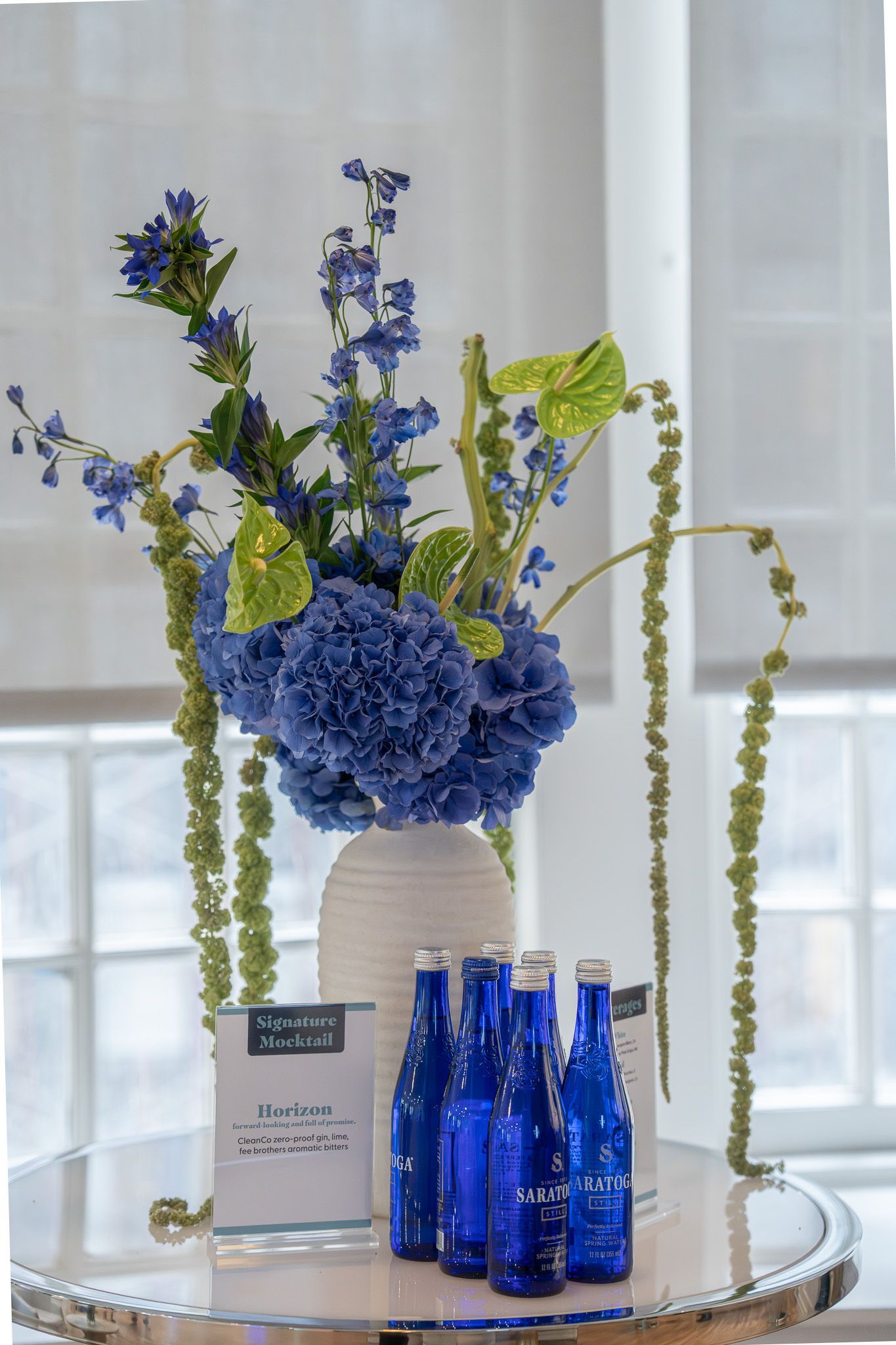 Blue and green floral arrangement on a table with blue bottles in front of a window