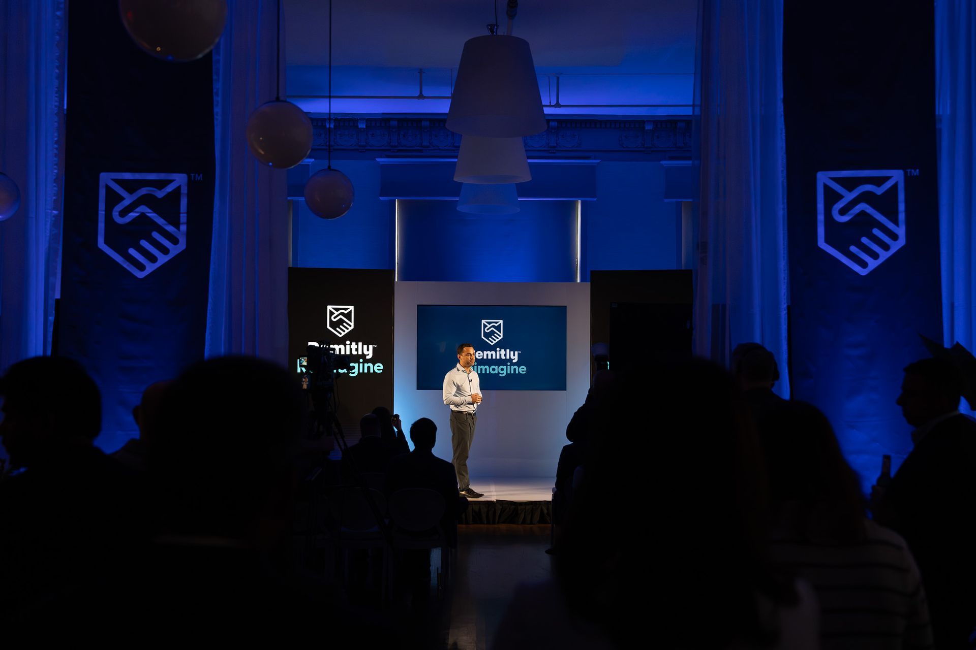 Person speaking on a blue-lit stage before an audience, with screens and logos behind them