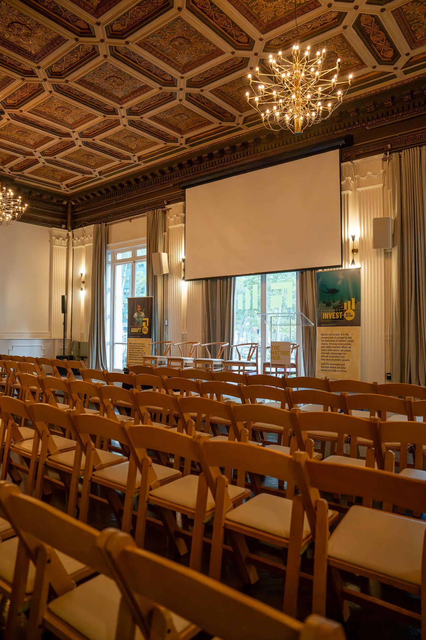 Empty conference room with rows of wooden chairs, a projector screen, and ornate ceiling chandeliers.
