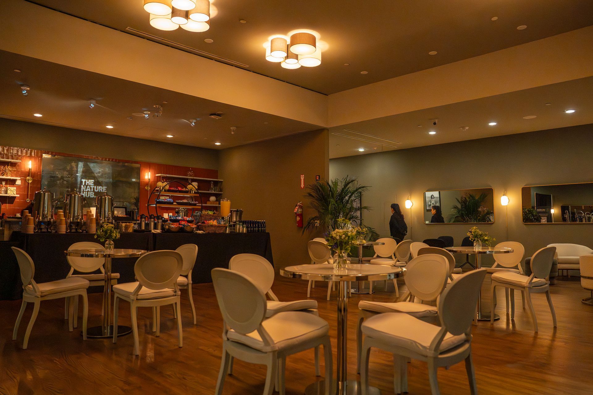 Warmly lit restaurant dining room with round tables, white chairs, and a bar in the background