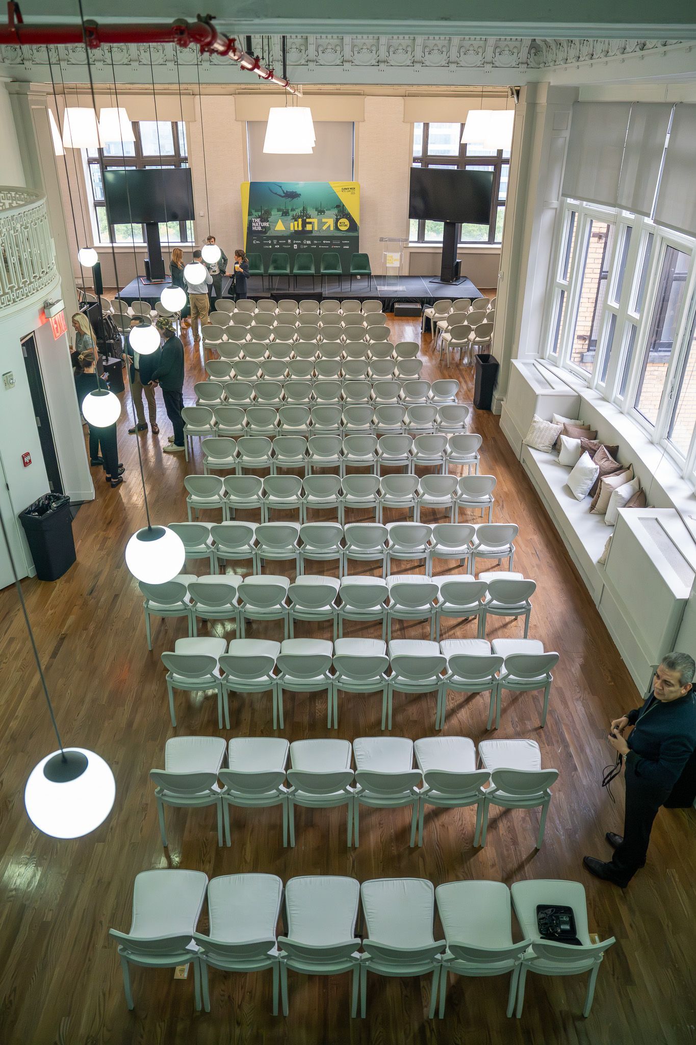 Rows of white chairs in a bright hall, with a few people standing along the sides.