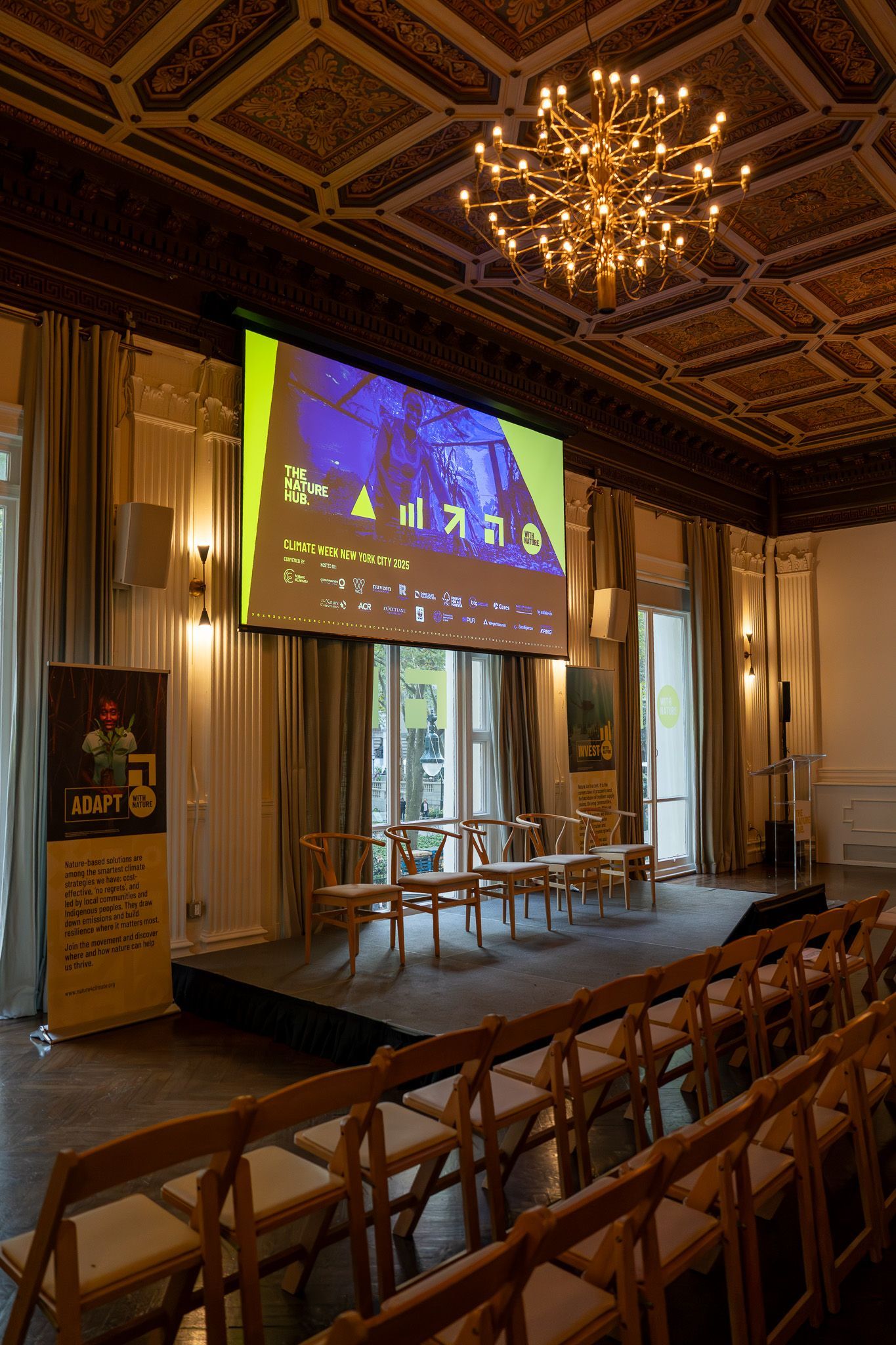 Elegant hall with stage, projector screen, rows of chairs, and ornate chandelier ceiling