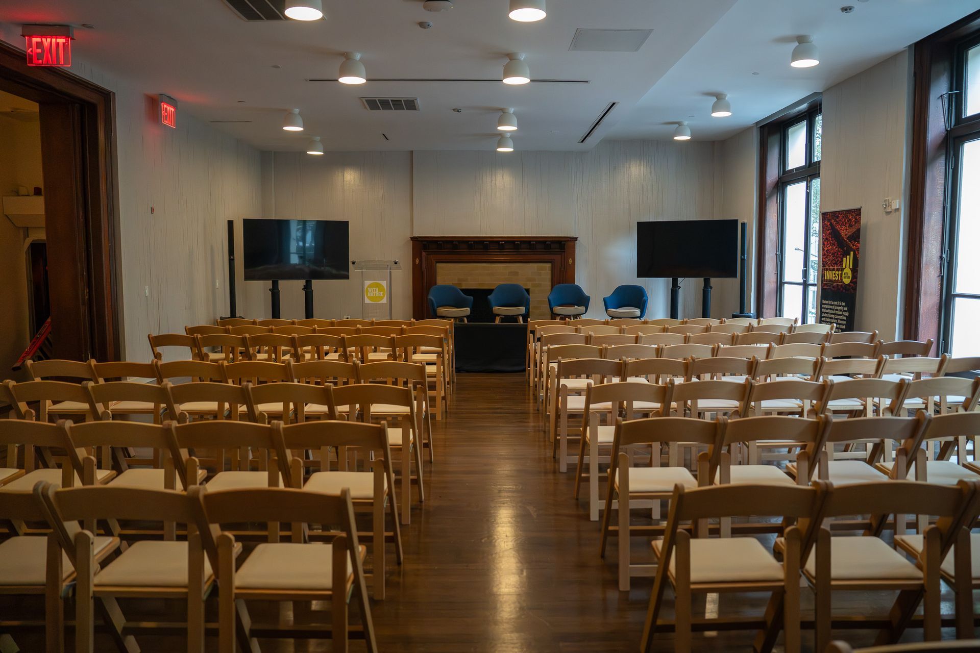 Empty conference room with rows of wooden chairs facing a front stage and screen.