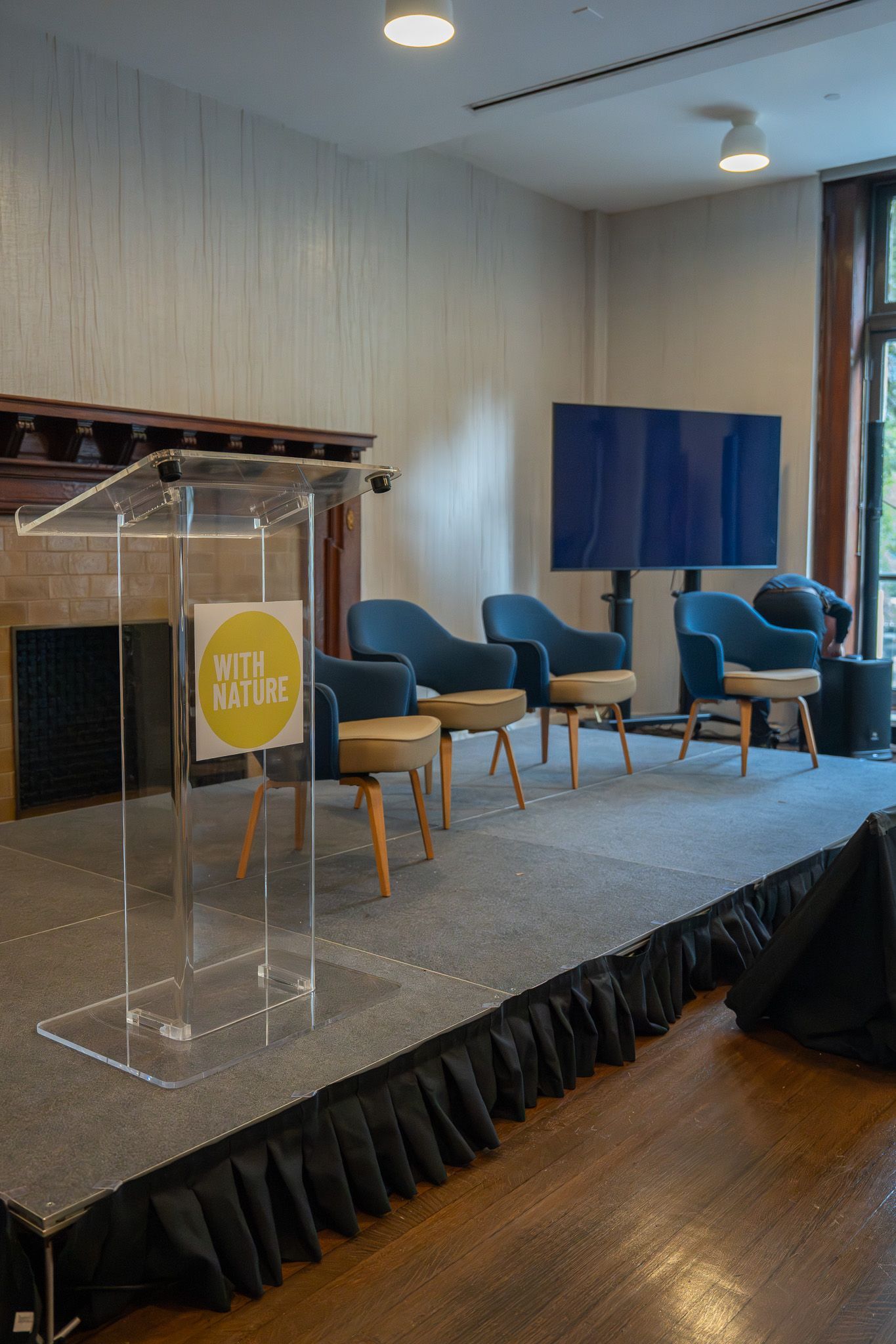 Empty stage with clear podium, blue chairs, and large blue backdrop in a room with wood floor.