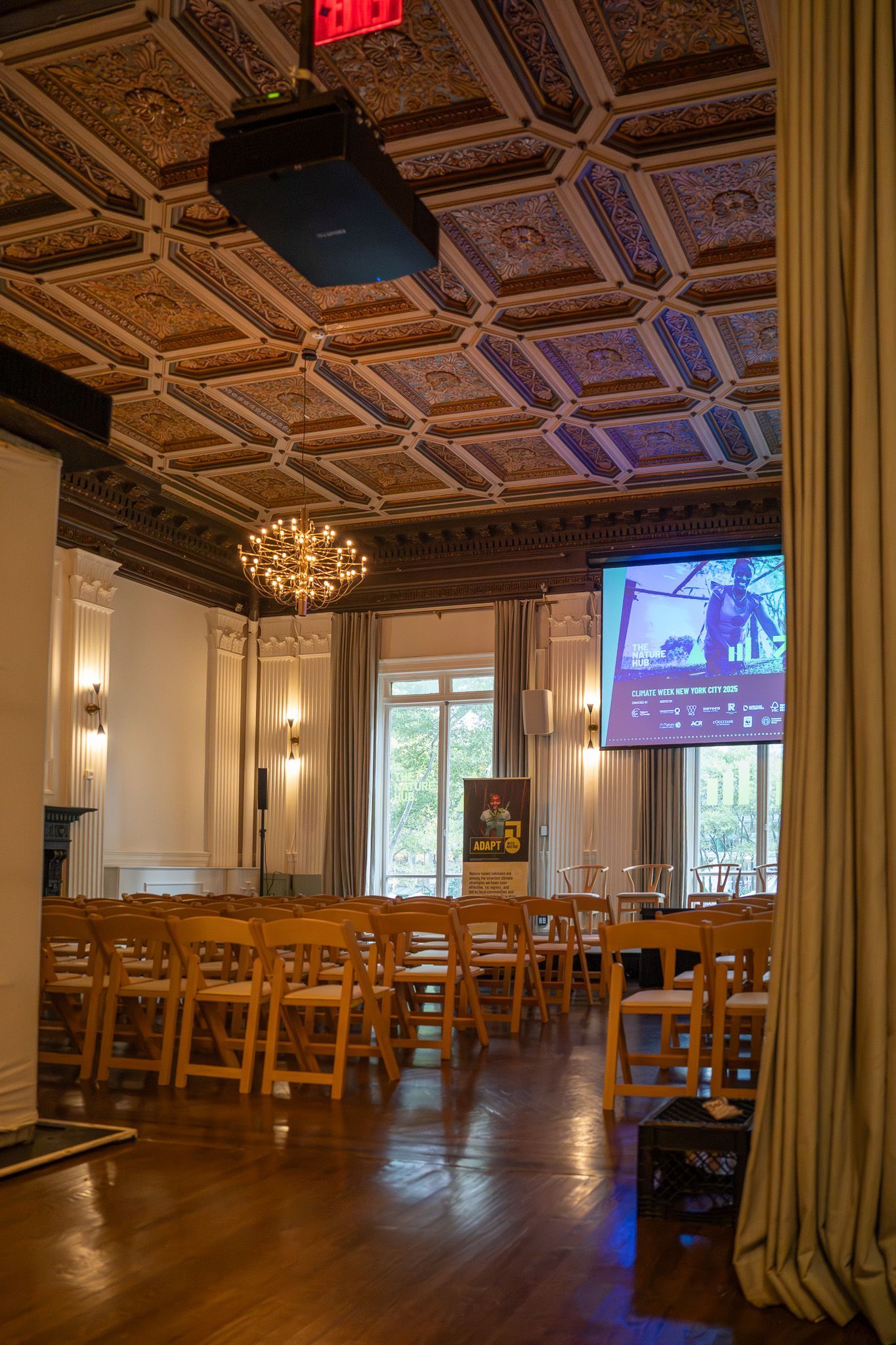 Elegant hall with wooden chairs, ornate ceiling, chandelier, and a projected screen near tall windows.