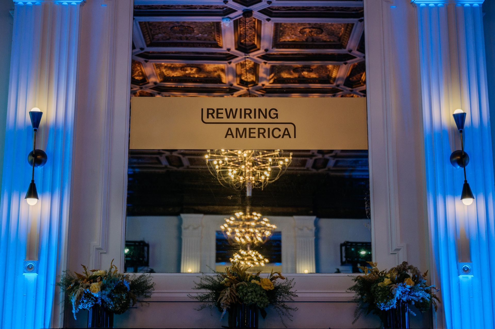 “Rewiring America” sign above a blue-lit lobby with a hanging chandelier and plants