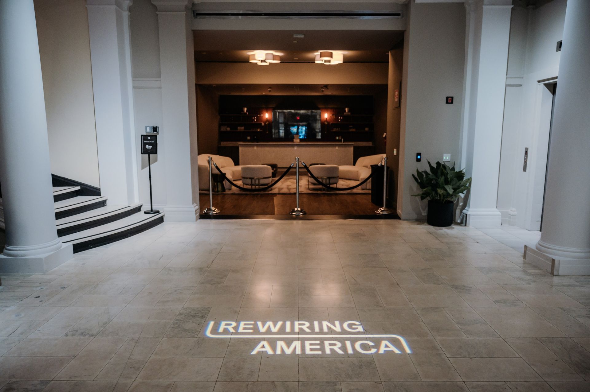 Lobby with large “REWIRING AMERICA” projection on the floor, leading to a warmly lit seating area.