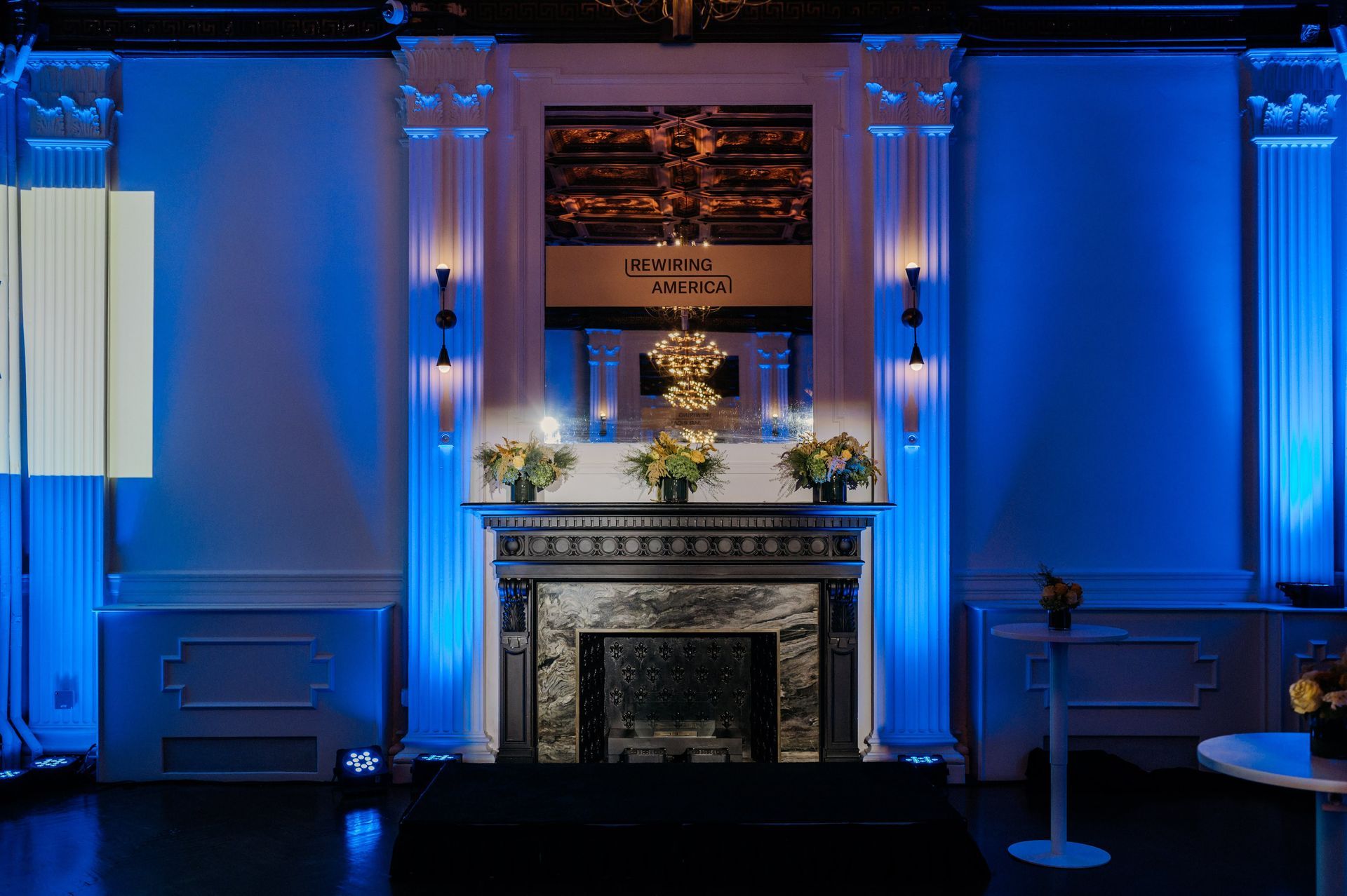 Blue-lit room with a fireplace, floral arrangement, and framed artwork on the mantel.