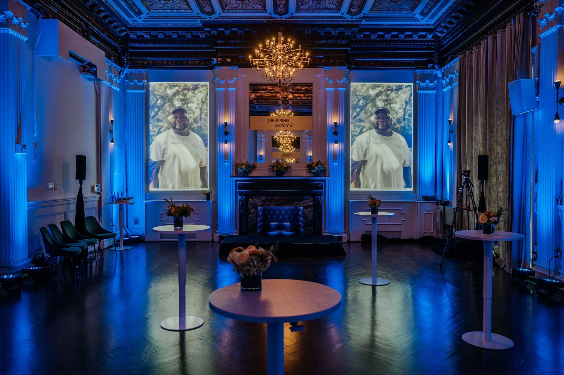 Blue-lit ornate room with two projected portraits on screens, tables, and chandeliers