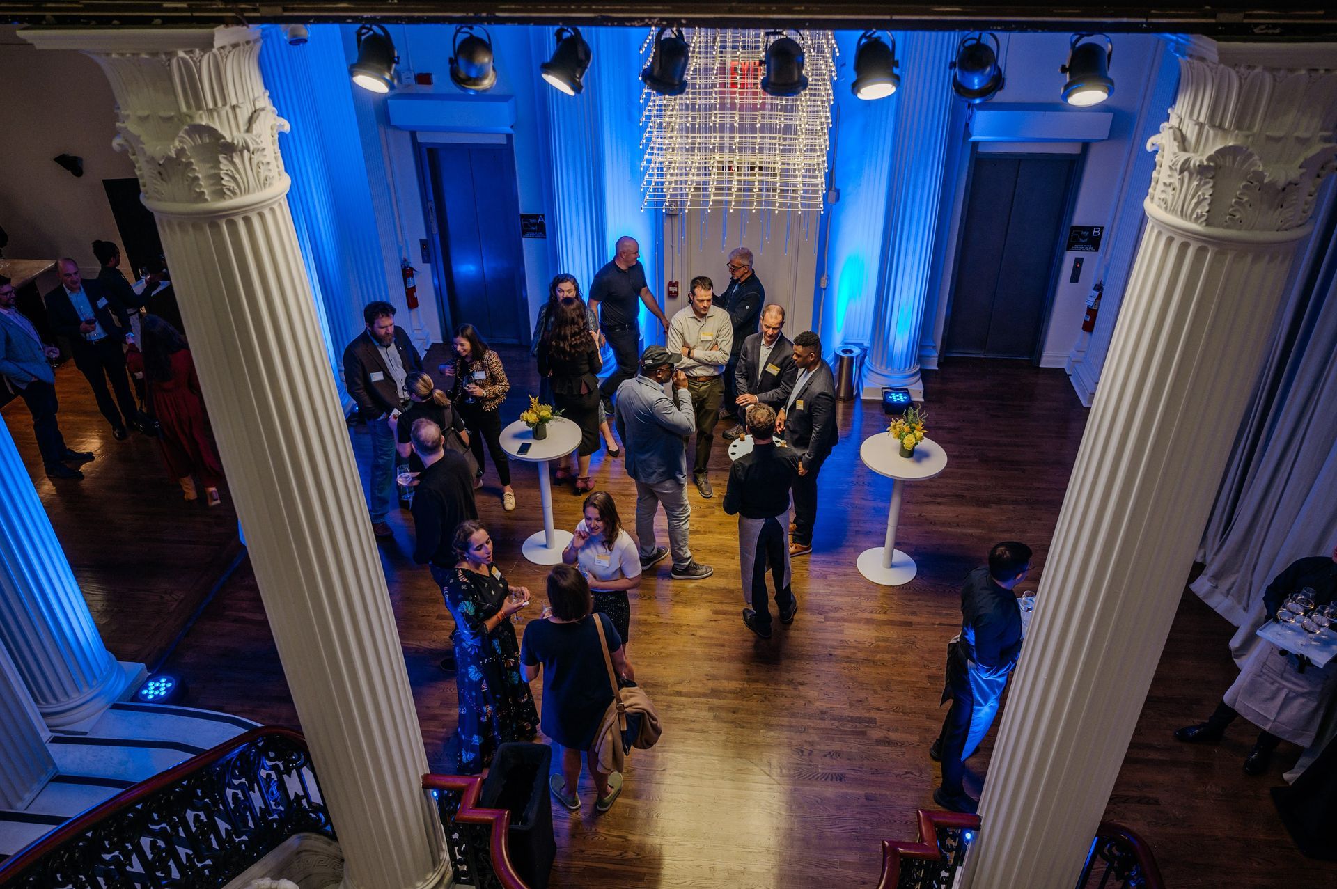 Guests mingling in a blue-lit hall with white columns and wooden floors, viewed from above