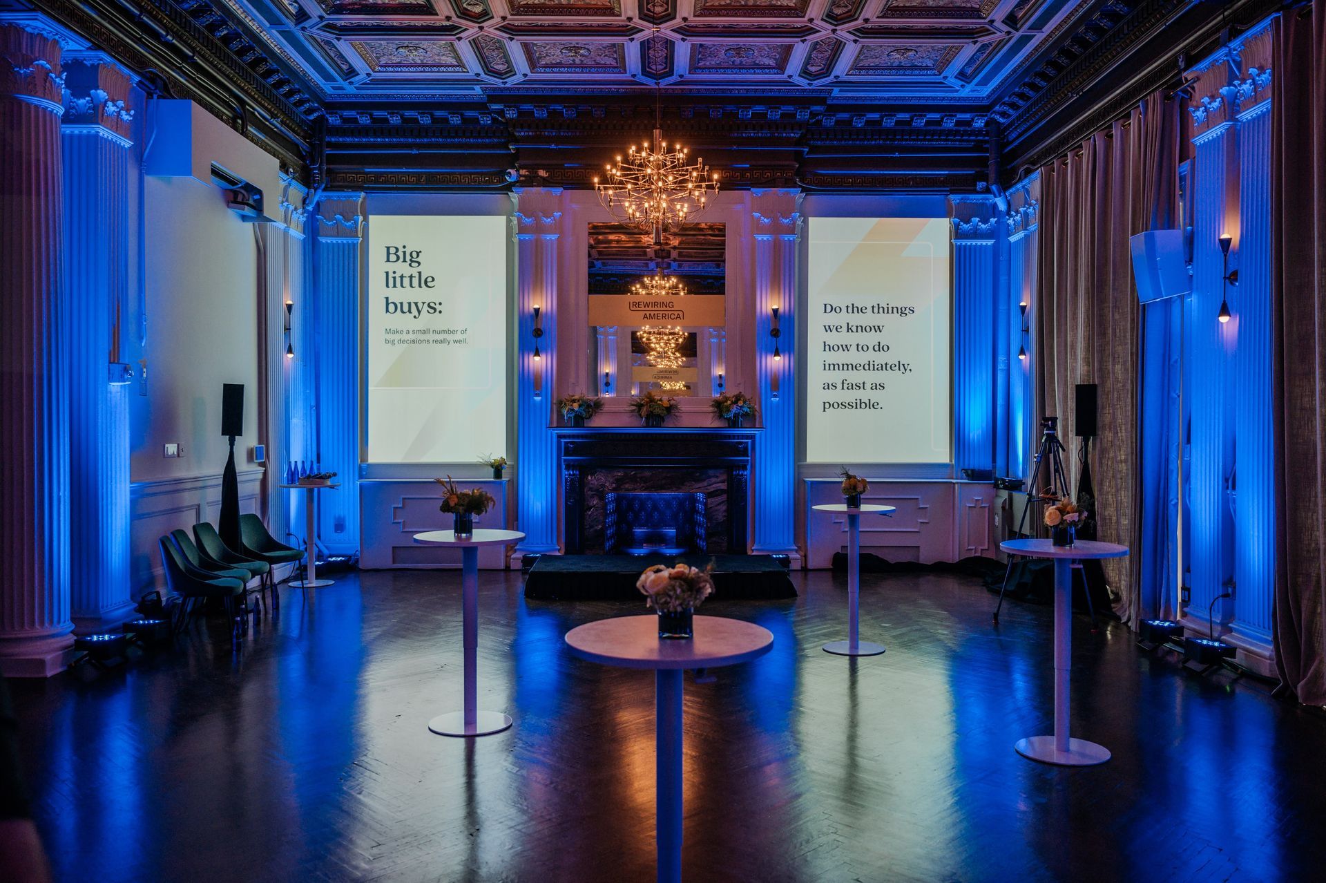 Blue-lit event hall with ornate ceiling, two projection screens, and cocktail tables in a formal setting