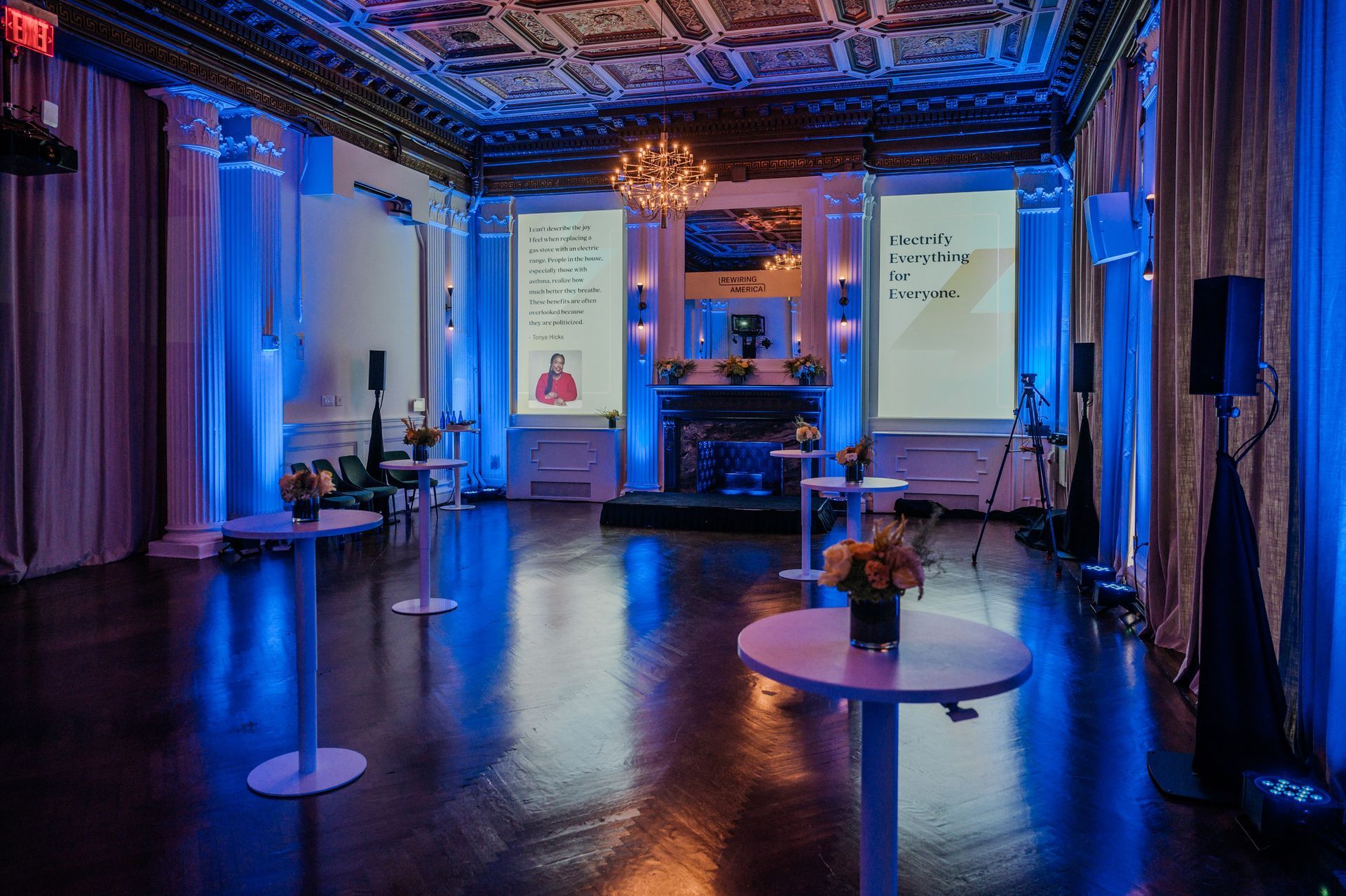 Blue-lit presentation room with two projection screens, tables, and chairs in an ornate venue