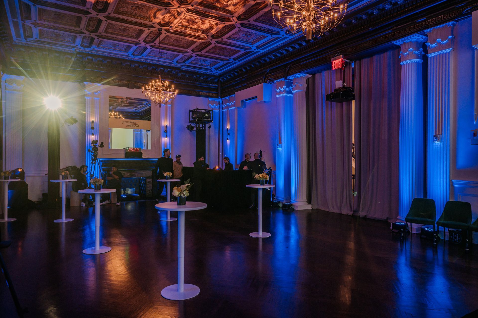 Elegant event hall with blue uplighting, chandeliers, and cocktail tables on a dark polished floor