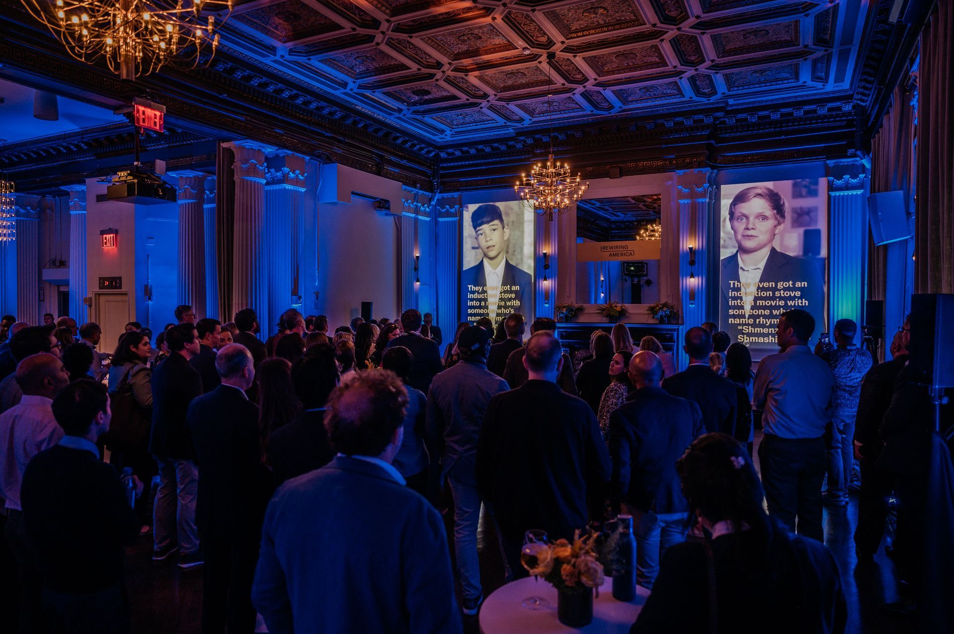 Crowded conference hall with blue lighting and large stage screens showing a speaker and presentation