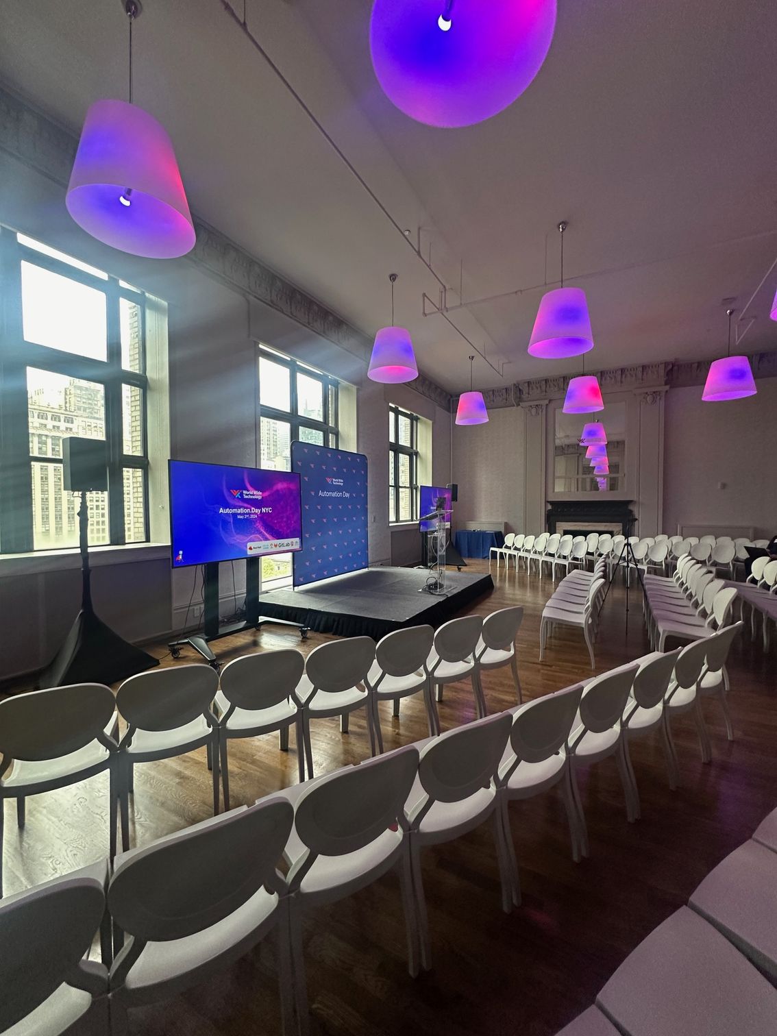 Conference hall with rows of white chairs, a stage, and purple hanging lights by large windows.