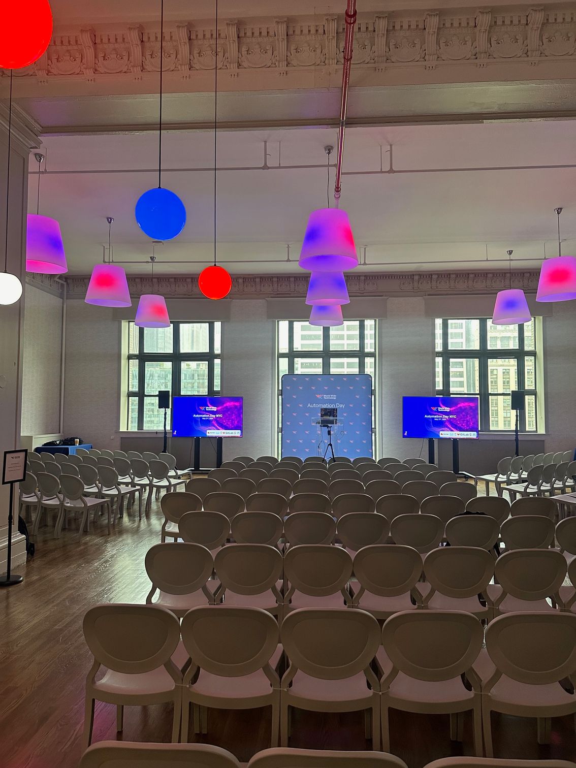 Rows of chairs in a decorated hall with hanging pink, blue, and red lights facing a stage and screens
