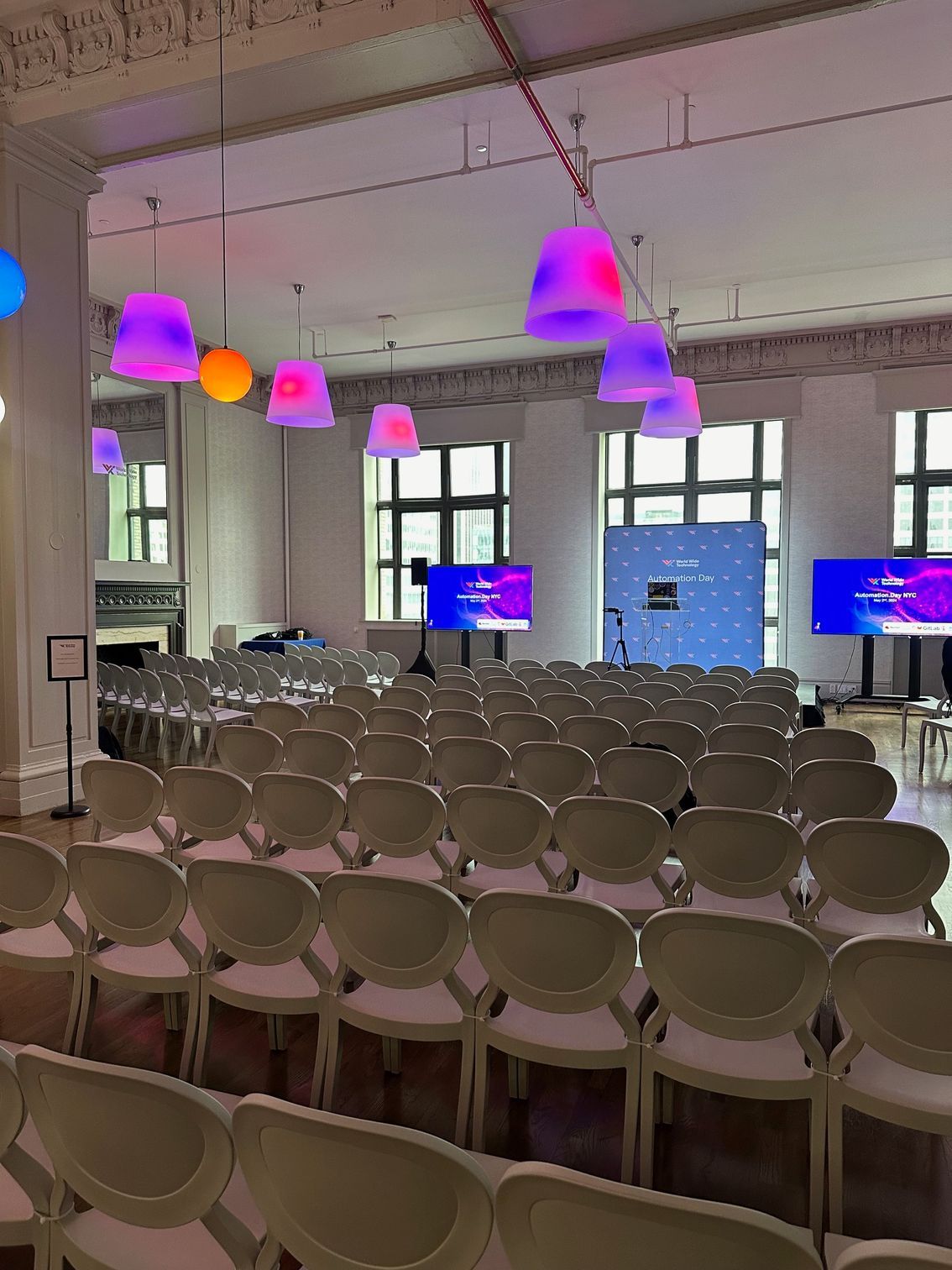 Rows of empty chairs in a bright event room with hanging purple and orange pendant lights and screens at the front