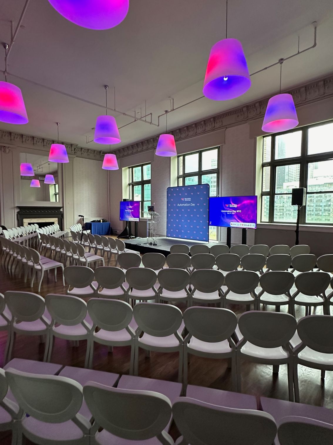 Empty conference room with rows of chairs and purple pendant lights, front screens lit near large windows