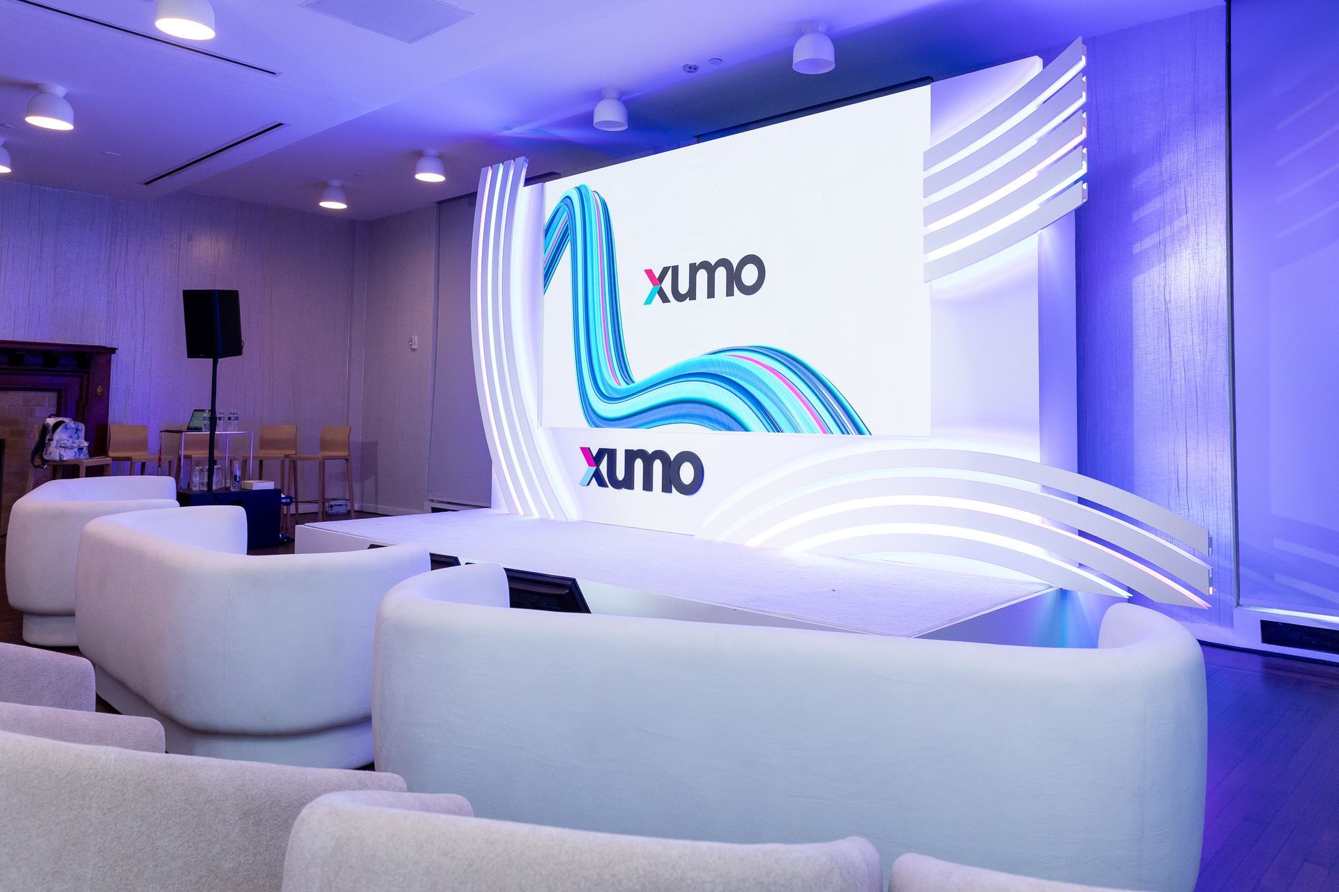 Modern event room with white sofas, blue lighting, and a large Xumo logo on a stage screen