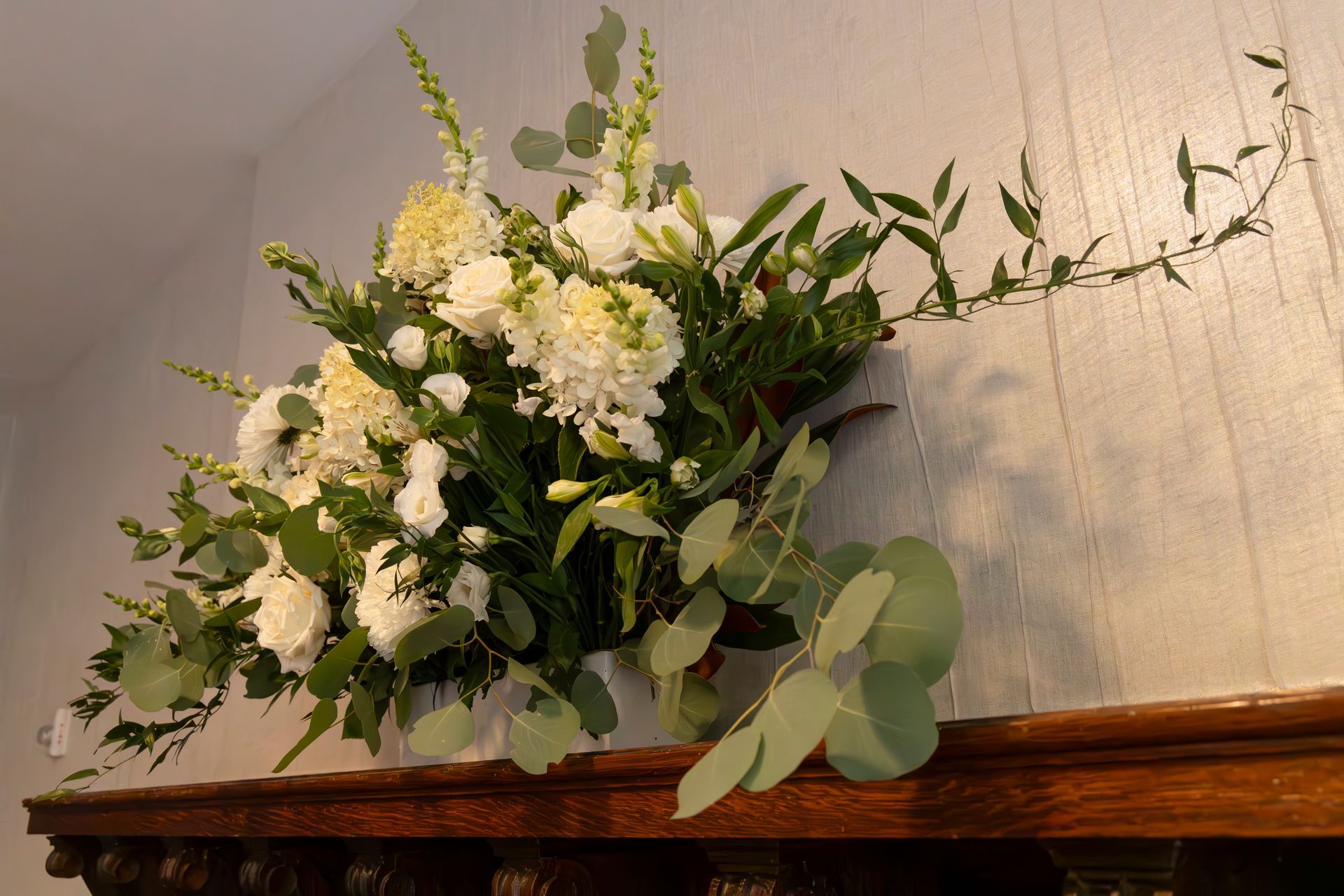 White floral arrangement with greenery displayed on a wooden mantel against a beige wall