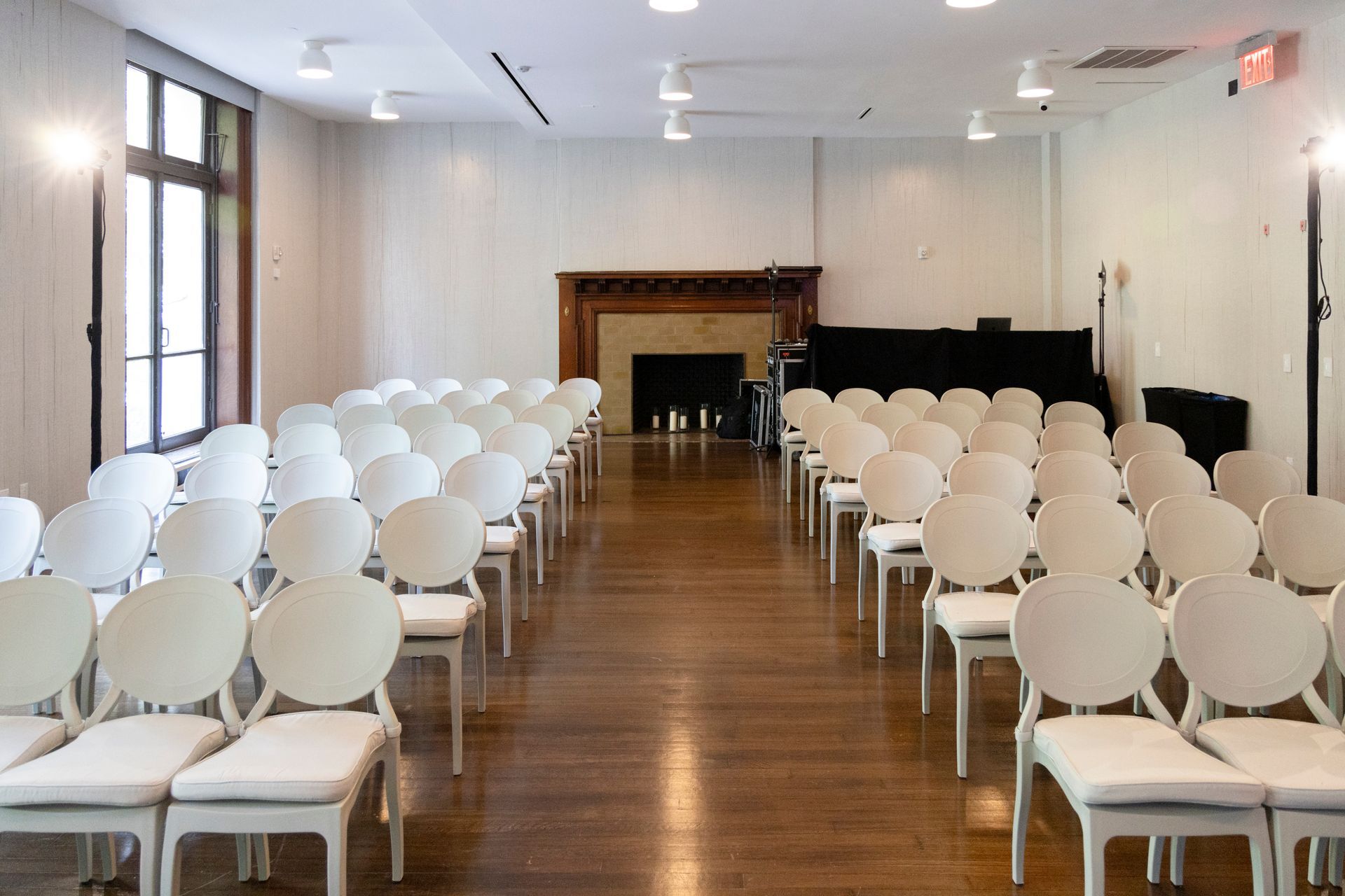 Rows of white chairs in a bright room with wooden floors and a fireplace at the front