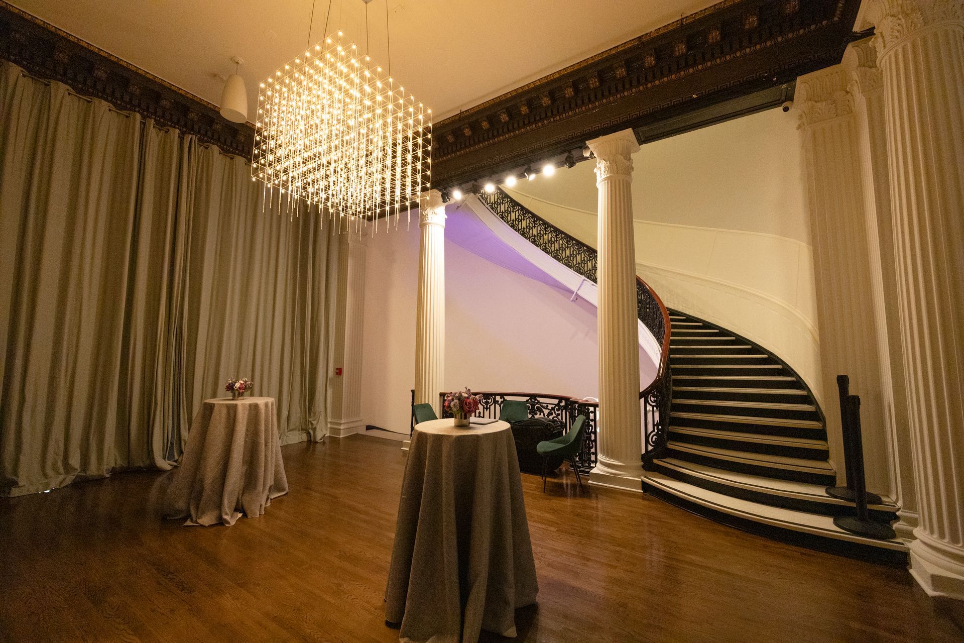Elegant ballroom with chandelier, draped curtains, cocktail tables, and a curved staircase